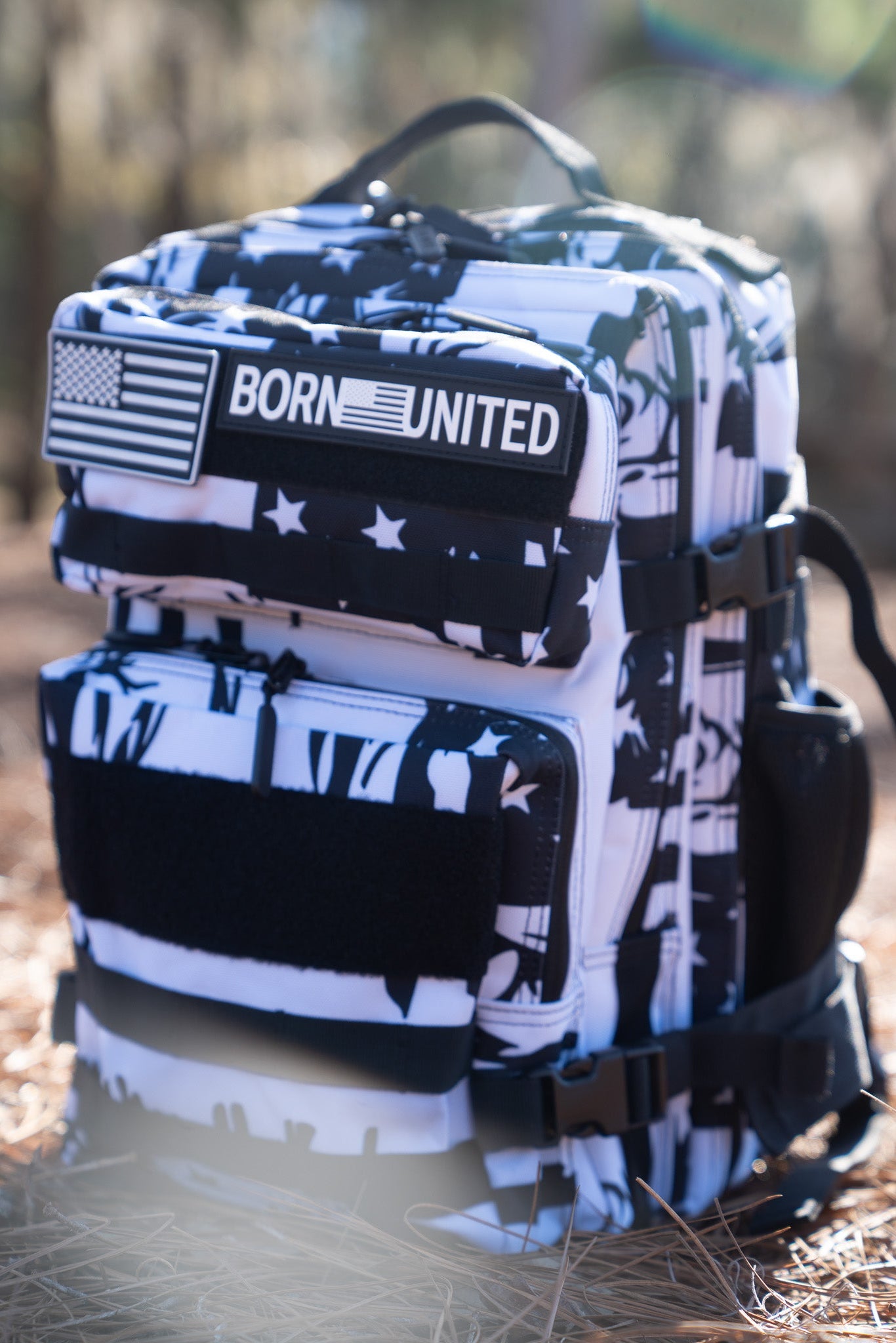 The American Stealth Special Edition Tac-Pac - 25L by Born United Private Label embodies American fashion with its black and white abstract design adorned with stars and a BORN UNITED patch. It rests on dried grass, with blurred trees in the background, echoing stealth clothing essence.