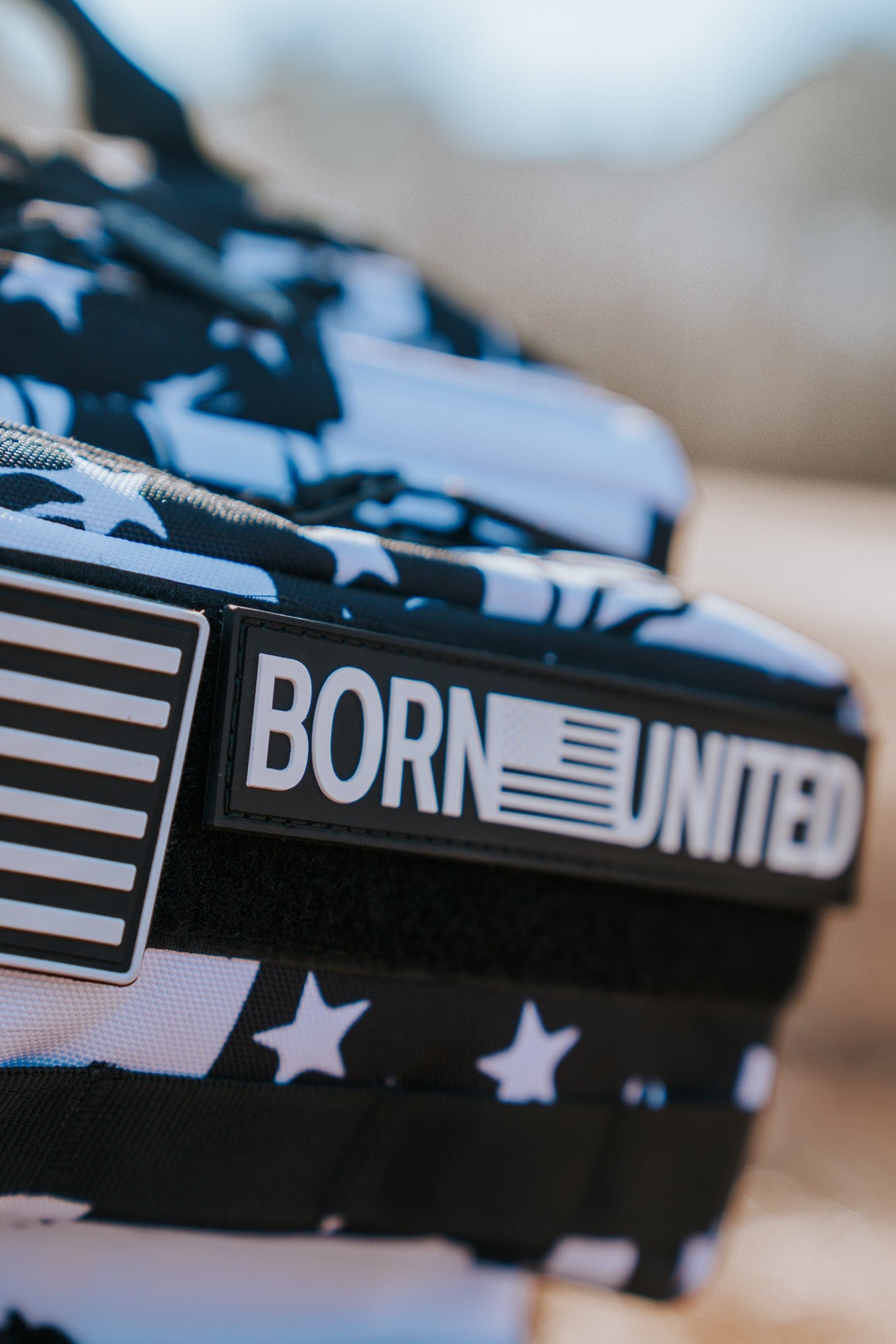 Close-up of the American Stealth Special Edition Tac-Pac - 25L by Born United Private Label. It features a black-and-white American flag pattern with a BORN UNITED patch and an emblem, showcased against a blurred outdoor background.