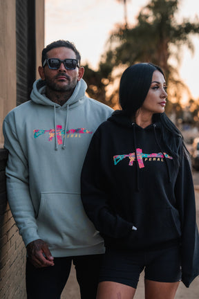 A man and woman wearing the Feral - Premium Dusty Sage Hoodie, printed with a colorful floral rifle graphic and the word FERAL, embodying a sense of radical authenticity.
