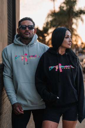 A man and a woman stand side by side wearing the Feral - Premium Hoodie by Tango Charlie Apparel, which features a floral-patterned rifle graphic and the word FERAL.