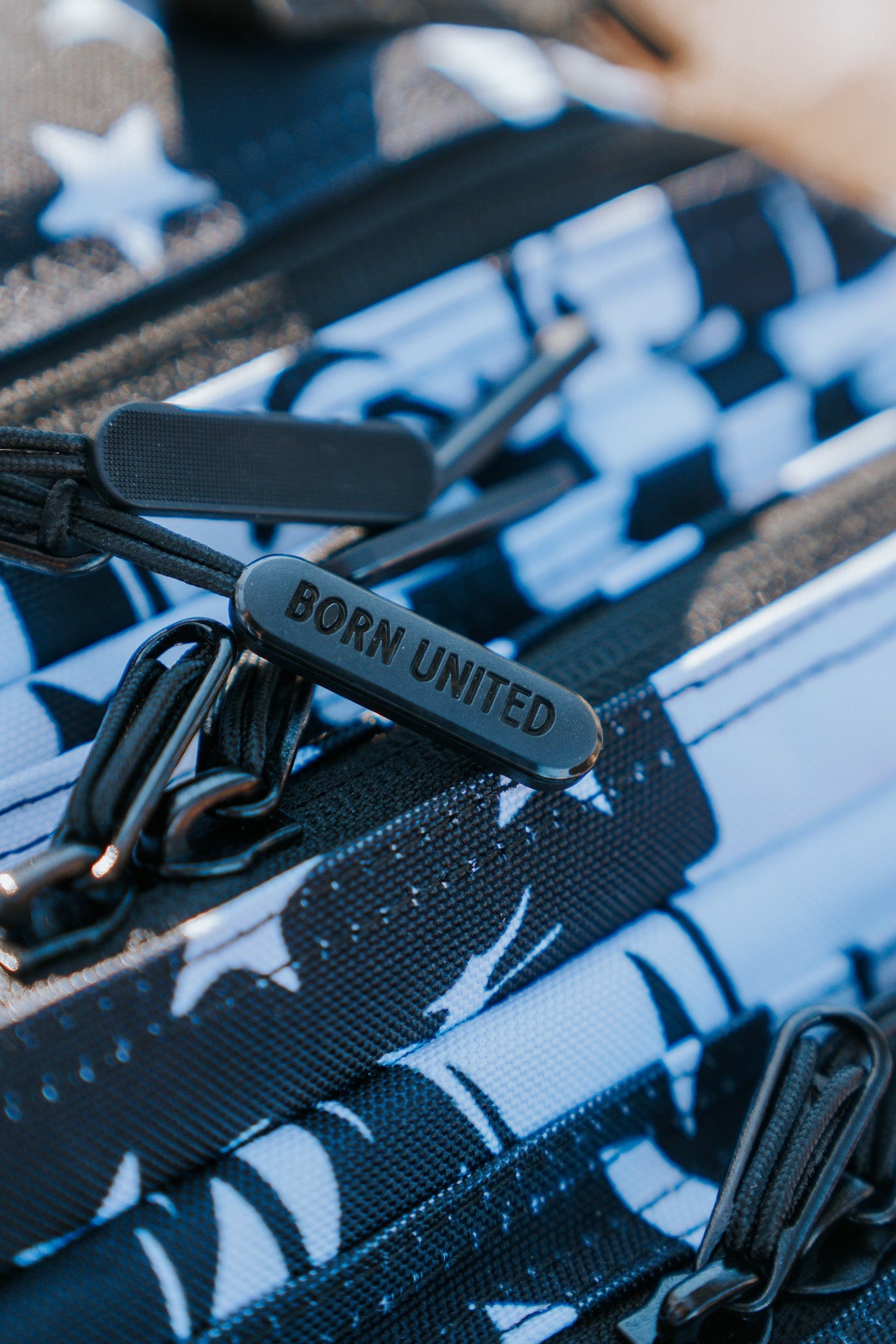 Close-up of a black zipper pull on the American Stealth Special Edition Tac-Pac - 25L by Born United Private Label. This backpack features a bold yet understated geometric pattern in black, white, and blue with star designs and multiple zippers, capturing the essence of modern American fashion.