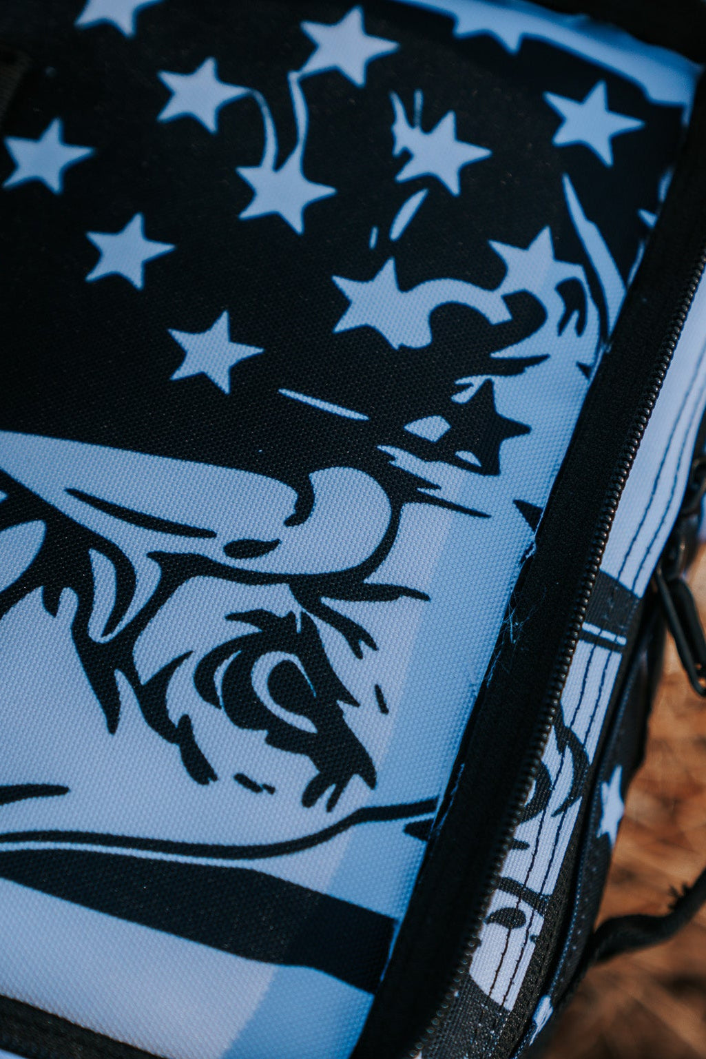 Close-up of an American Stealth Special Edition Tac-Pac - 25L from Born United Private Label, featuring bold black and white graphics with eye and head motifs. Partly zipped, it sits on a straw-like surface under bright lighting, exuding the intriguing world of stealth clothing.