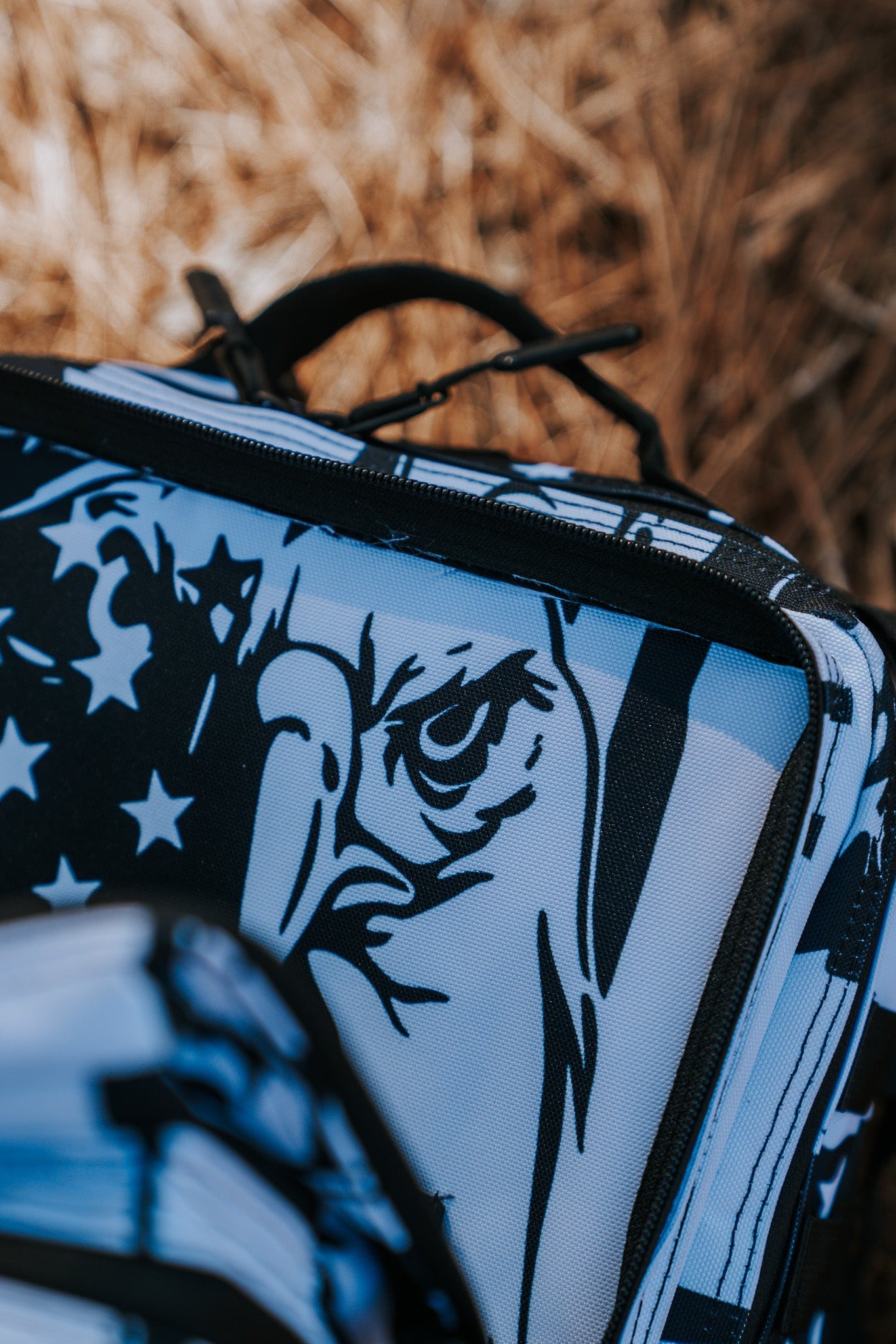 A close-up of the Born United Private Labels American Stealth Special Edition Tac-Pac - 25L features a stylized eagle and stars in black and white, capturing American fashions essence. The dry grass background hints at an outdoor setting where this stealth gear blends into nature seamlessly.