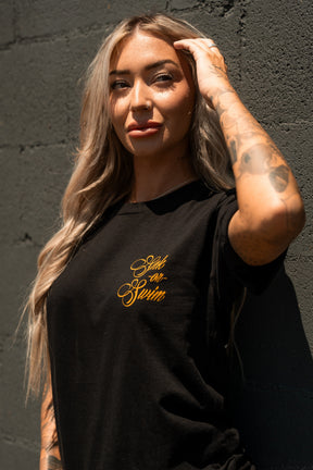 A woman with long blonde hair and tattoos poses against a dark wall, wearing the Relentless Betrayal Sink Or Swim Kraken T-Shirt. She rests one hand on her head and confidently gazes at the camera.