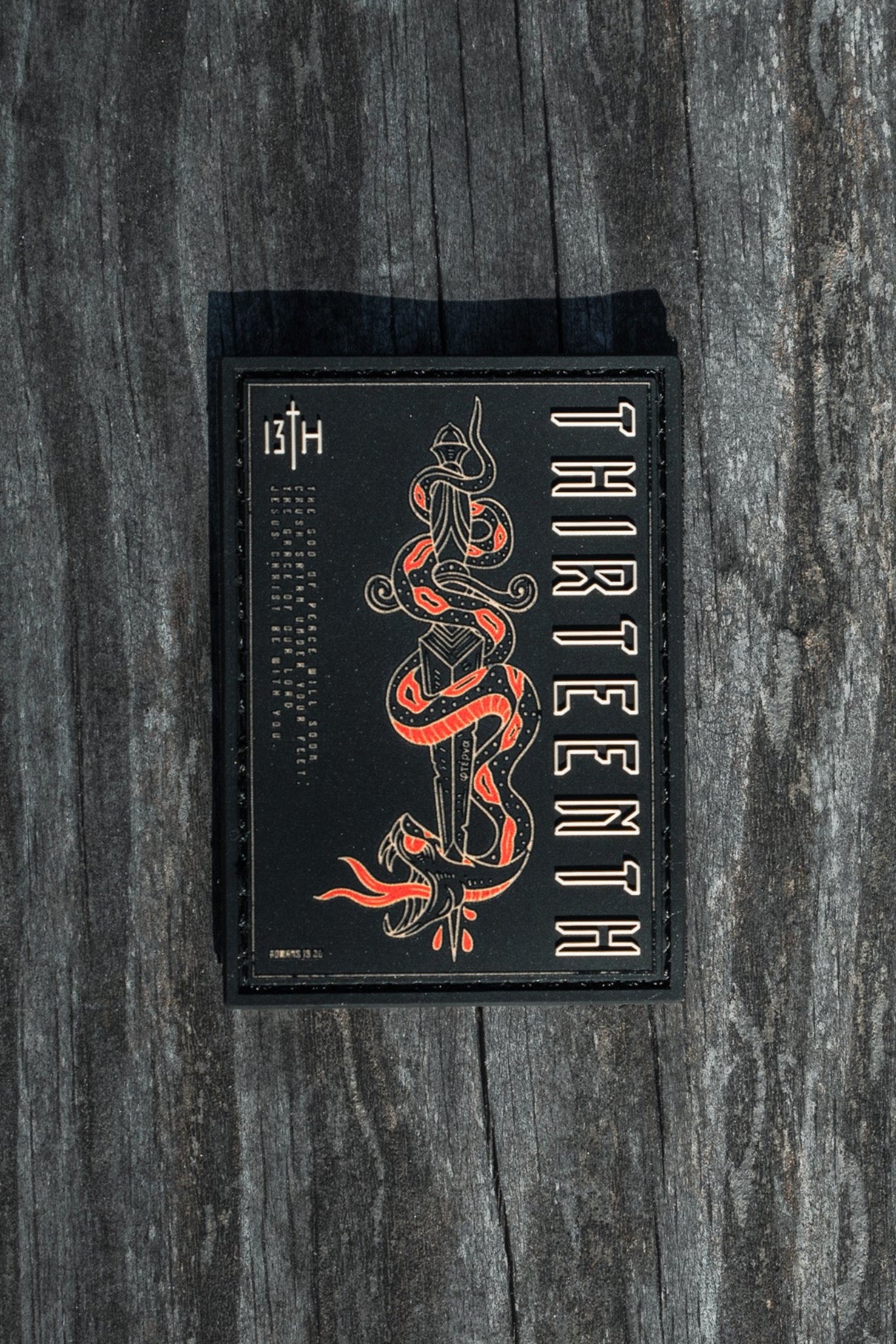 The product, Lucifers Demise PVC Patch by 13th, features a sleek black card with red and white graphics of a coiled snake and the word THIRTEENTH showcased vertically, beautifully set against a textured wooden background.