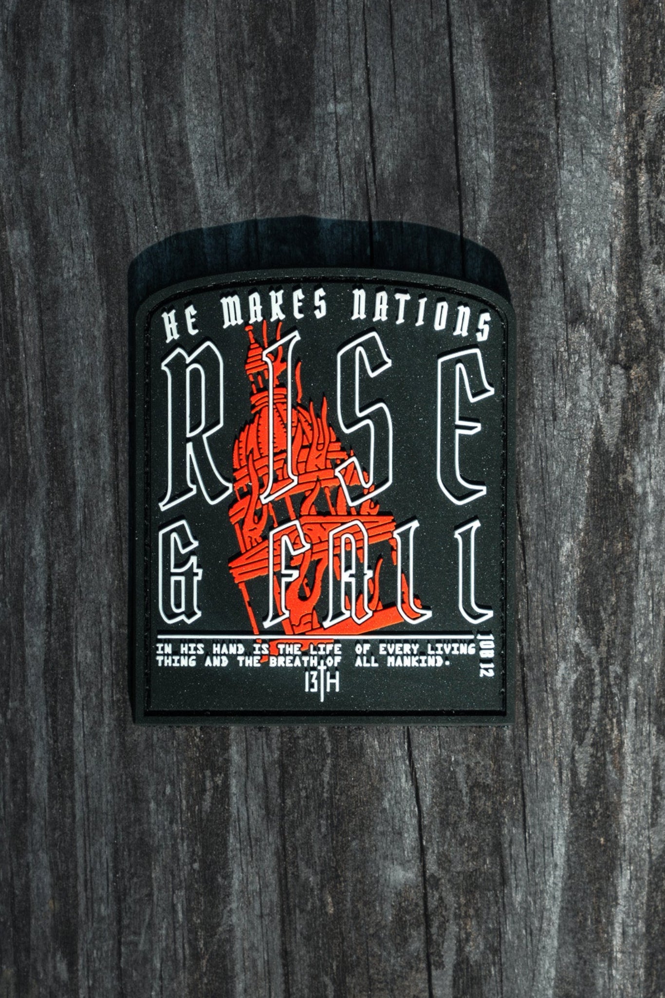 A Rise And Fall PVC Patch on wood features a red sword graphic. The text reads, He makes nations rise & fall. The life hand is in the hands of every living thing and the breath of all mankind. Brand: 13th, Reference: Thirteenth 13:14.