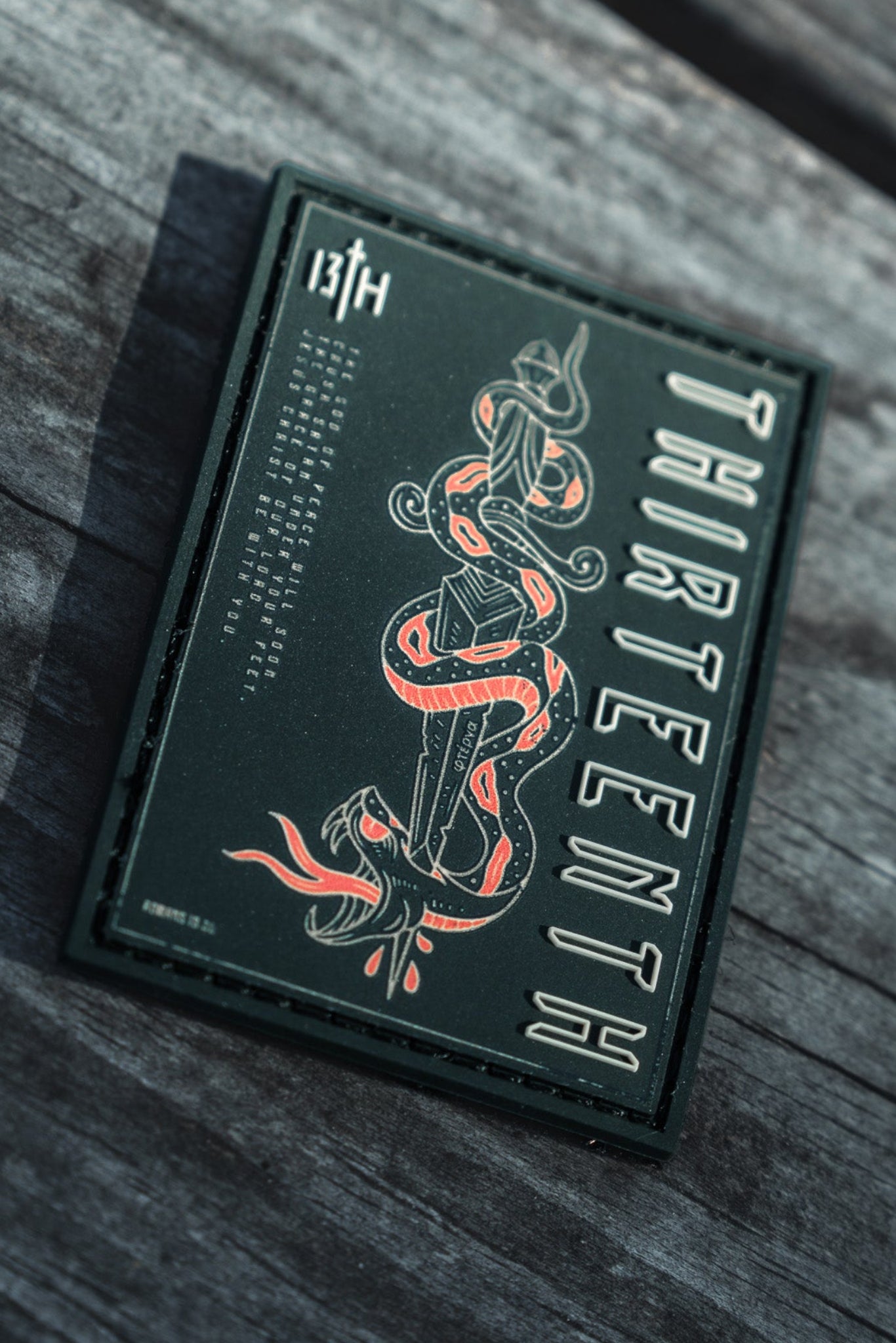 A dark green card showcases the 13th brand vertically and 13TH in the top corner, featuring a striking red and white snake design at its center, known as the Lucifers Demise PVC Patch. Its elegantly displayed on a wooden surface.