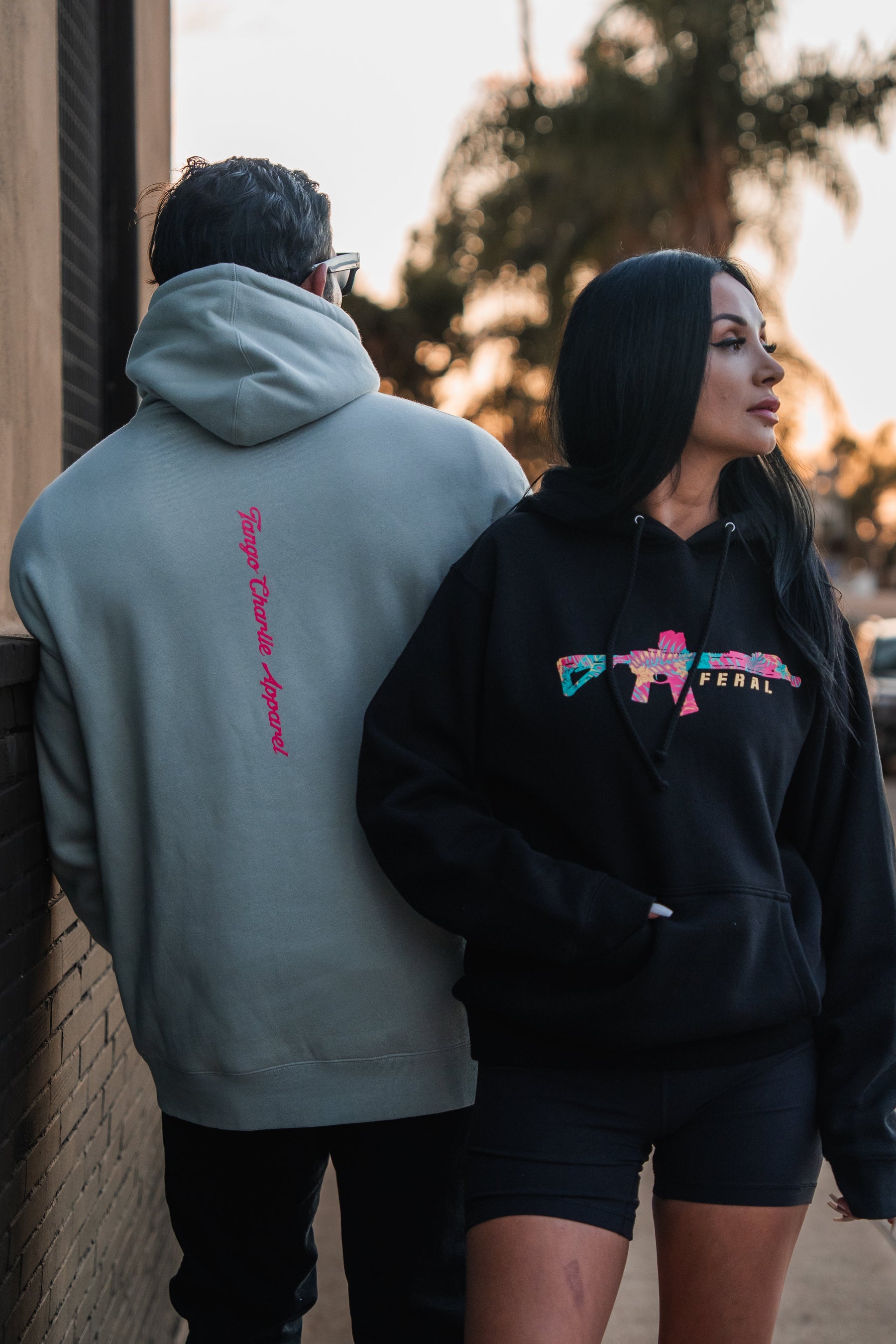 Two people stand back-to-back wearing Feral - Premium Hoodies, the person on the left in a light gray hoodie with pink text and the person on the right in a black graphic hoodie.
