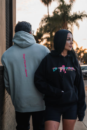A man wearing a Feral - Premium Dusty Sage Hoodie and a woman in a black hoodie with a colorful rifle graphic stand side by side, capturing an unfiltered observation of Feral Wild style.