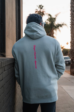 A person seen from behind wearing a Feral - Premium Dusty Sage Hoodie with Tango Charlie Apparel printed in red vertically down the center back, capturing a moment of radical authenticity.
