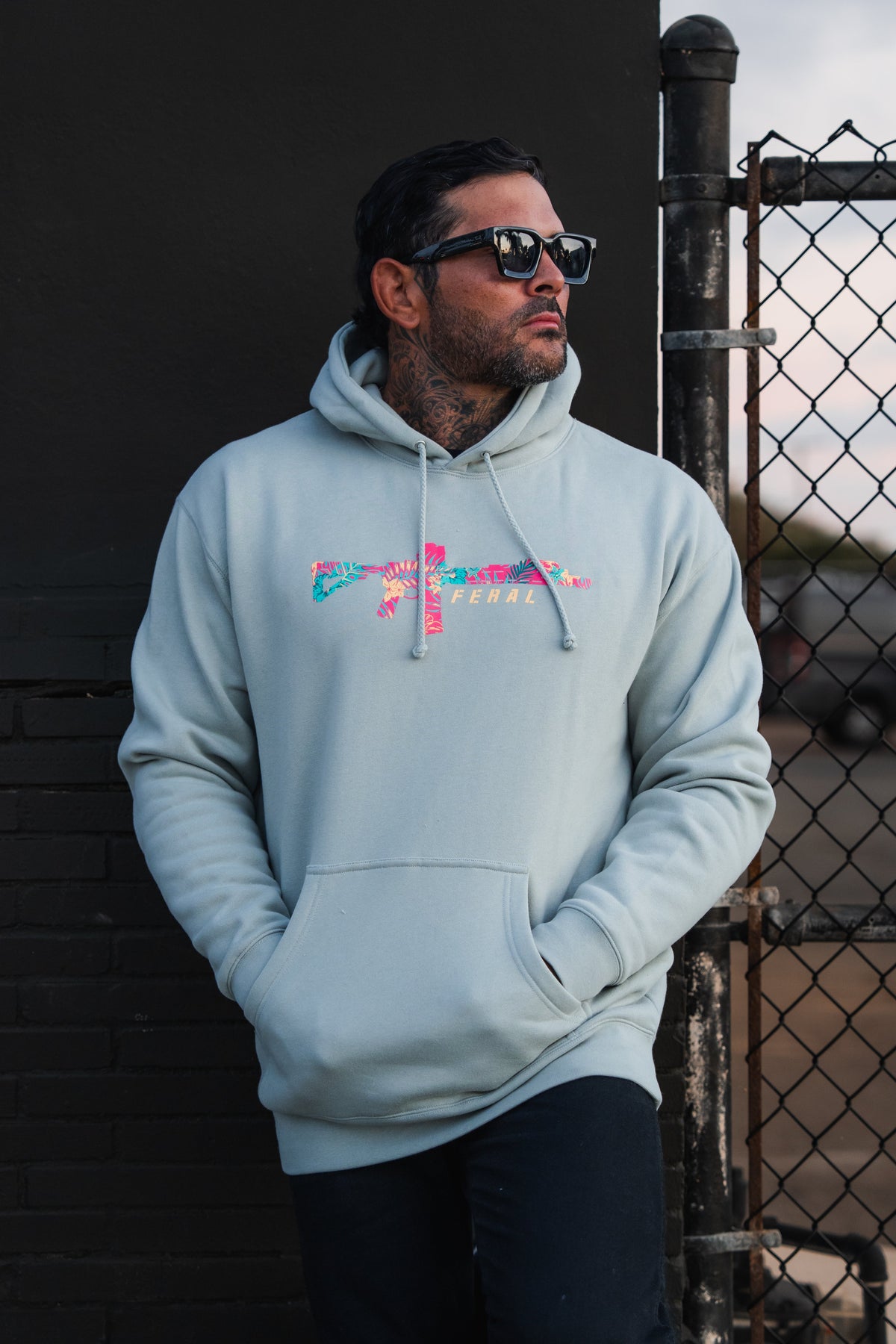 A man wears a Feral - Premium Dusty Sage Hoodie featuring a floral rifle graphic and the word FERAL.