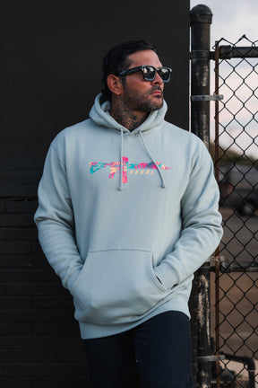 A man wears a Feral - Premium Dusty Sage Hoodie featuring a floral rifle graphic and the word FERAL.