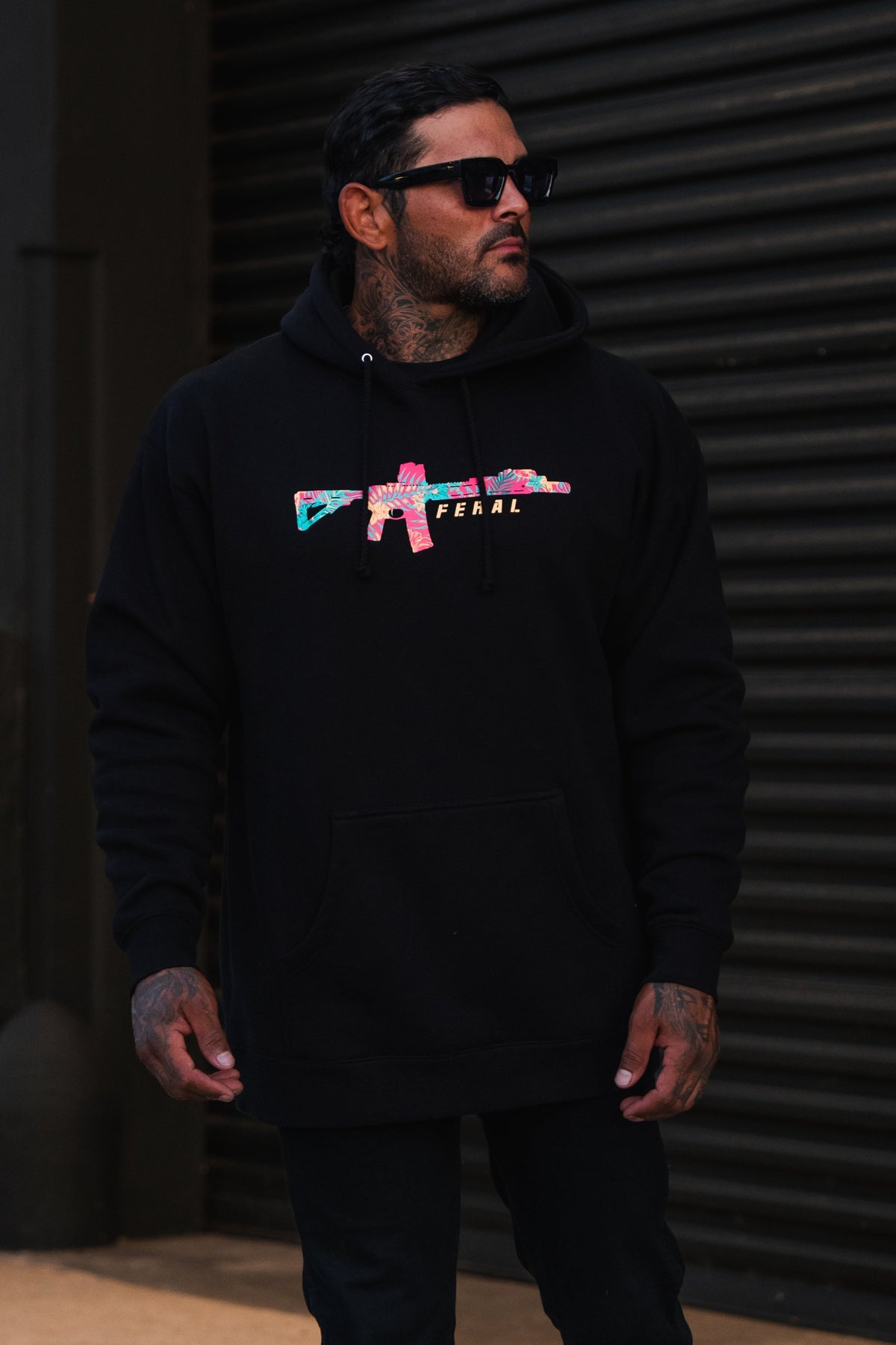 A man wears a black Feral - Premium Hoodie featuring a multicolored rifle graphic and the word FERAL, embodying a spirit of radical authenticity.