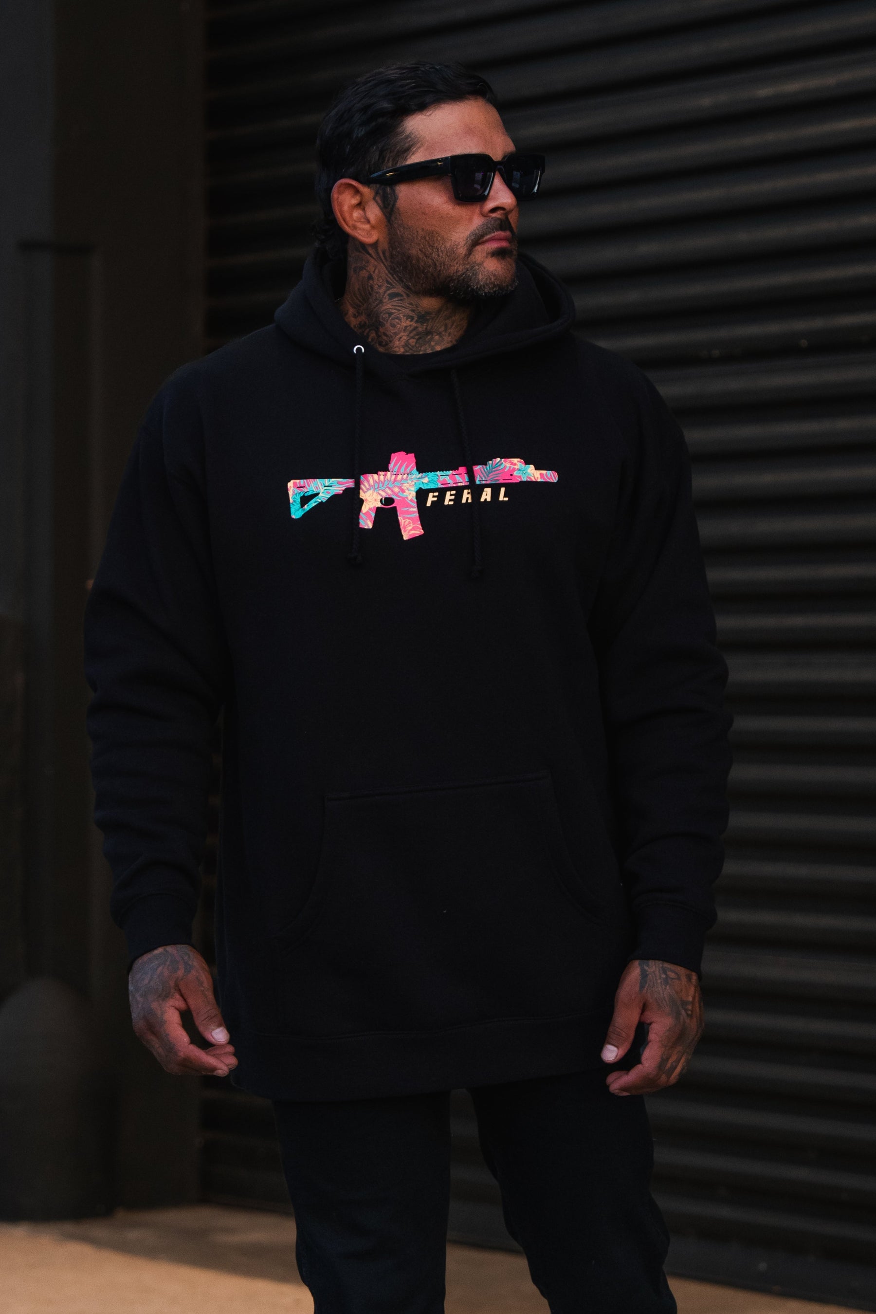 A man wears a black Feral - Premium Hoodie featuring a multicolored rifle graphic and the word FERAL, embodying a spirit of radical authenticity.