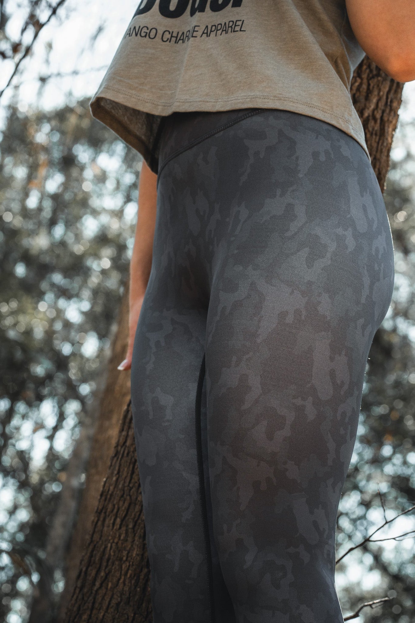 A person stands outdoors in a cropped shirt and Born United Private Labels Midnight Camo Leggings, with a tree trunk in the background, exuding an adventurous vibe typical of a wooded area.