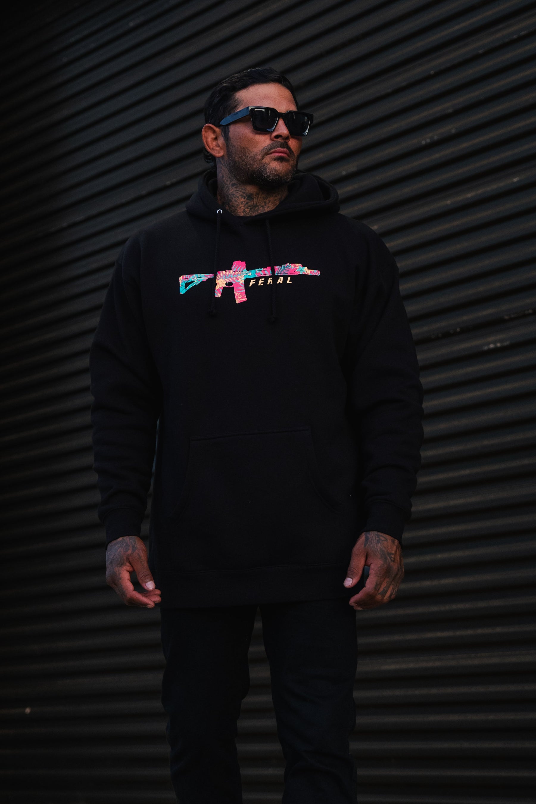 A man wears a black Feral - Premium Hoodie featuring a multicolored rifle graphic and the word FERAL.