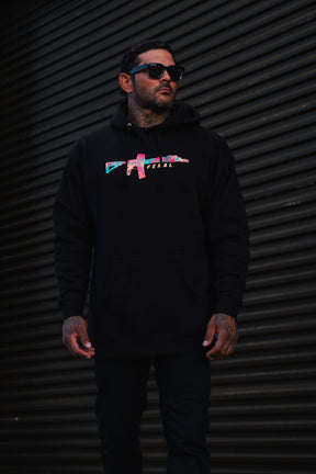 A man wears a black Feral - Premium Hoodie featuring a multicolored rifle graphic and the word FERAL.