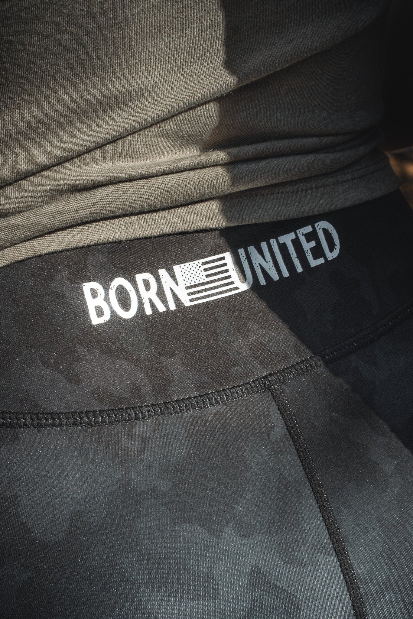 Close-up of the waistband on Midnight Camo Leggings by Born United Private Label, showcasing BORN UNITED text with an American flag. The individual is in a dark top.