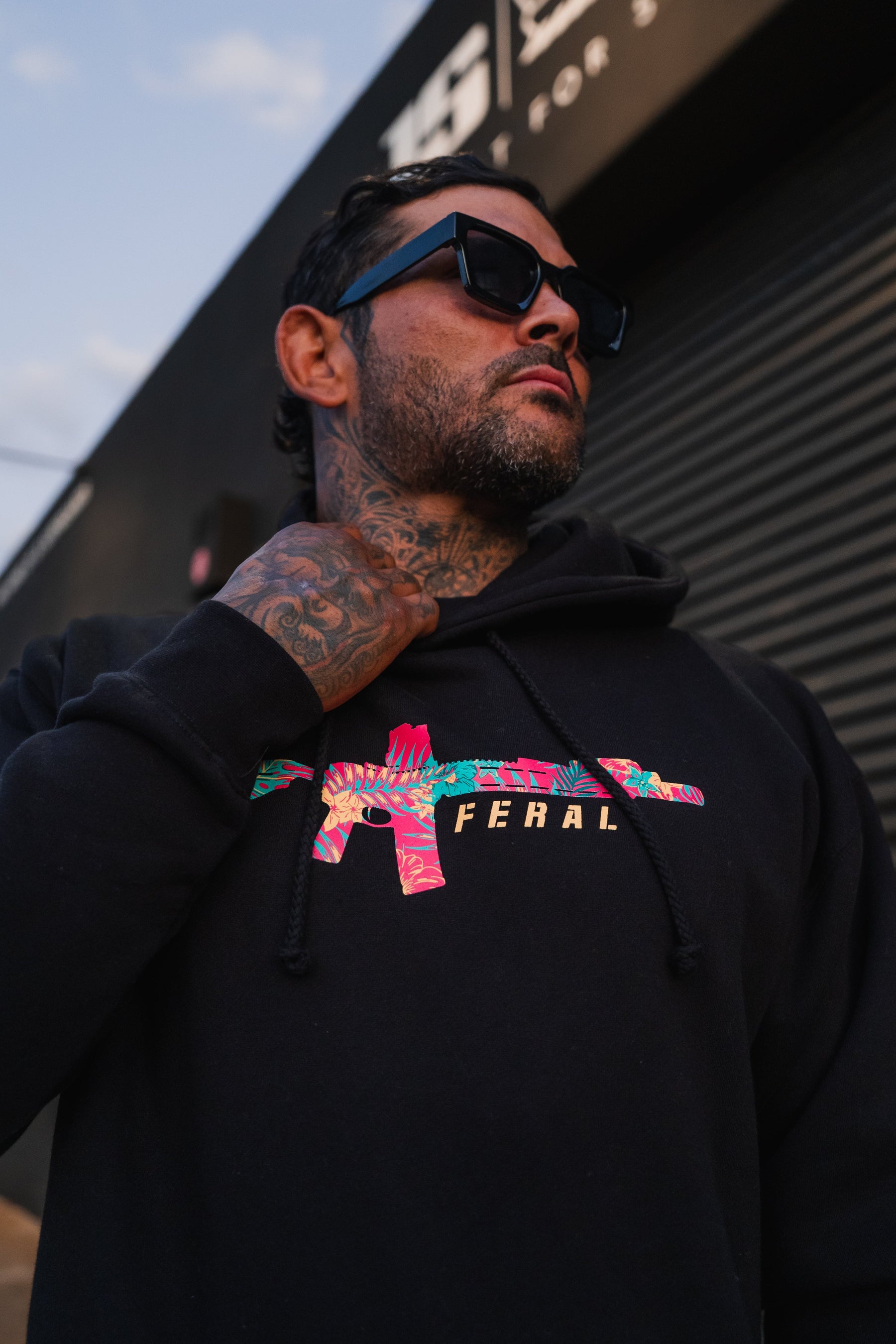 A man wearing a black Feral - Premium Hoodie featuring a tropical floral rifle graphic and FERAL text.