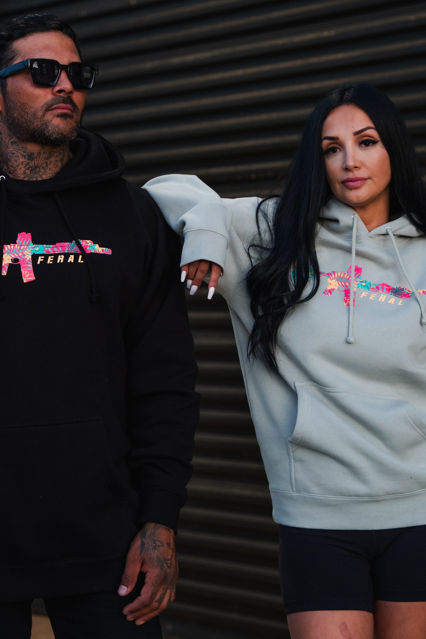 A woman in a Feral - Premium Dusty Sage Hoodie with a colorful rifle graphic leans on a man in a matching black hoodie, embodying radical authenticity in their unfiltered observation.