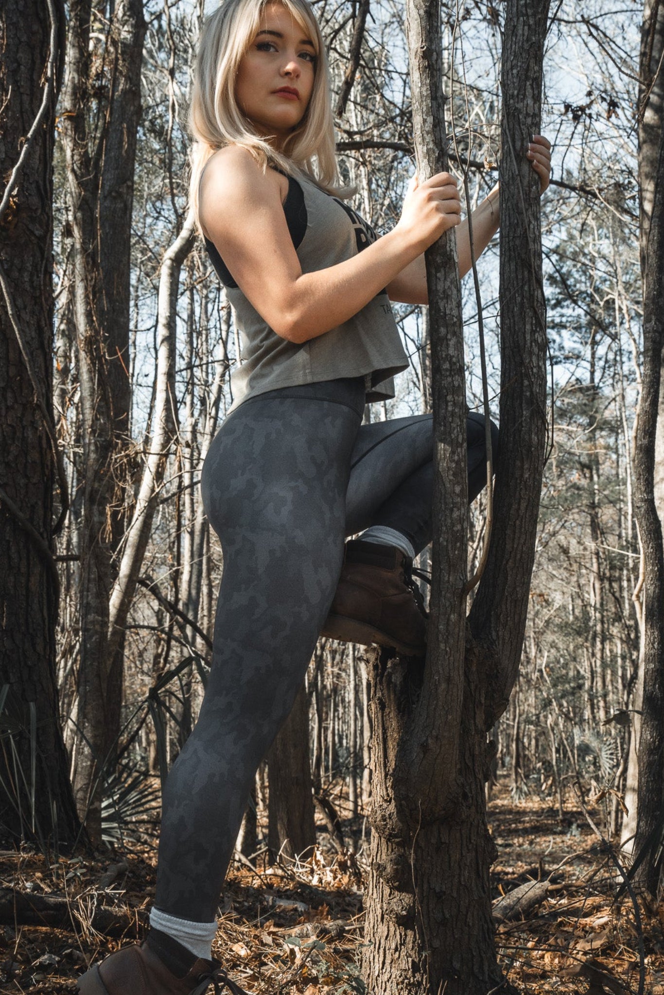 A platinum blonde woman climbs a tree in the forest, wearing a sleeveless top, brown boots, and Midnight Camo Leggings by Born United Private Label. Leafless trees fill the background, suggesting a winter or early spring setting.