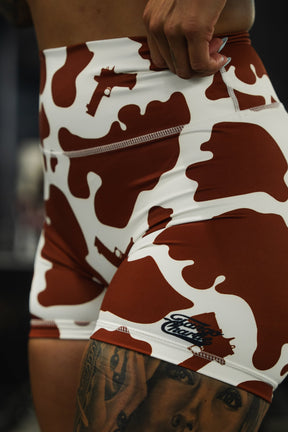 A close-up shows a person in Tango Charlie Apparel’s Women’s High-Waisted Seamless Front Compression Training Shorts in Moo Moo Pew cow print, with a tattooed leg visible and an indoor space blurred in the background.