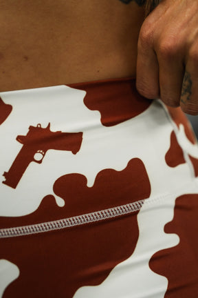 Close-up of a person’s hip wearing Tango Charlie Apparel's Moo Moo Pew Women’s High-Waisted Seamless Front Compression Training Shorts in brown and white camo with a gun graphic; a hand rests near the waistband.