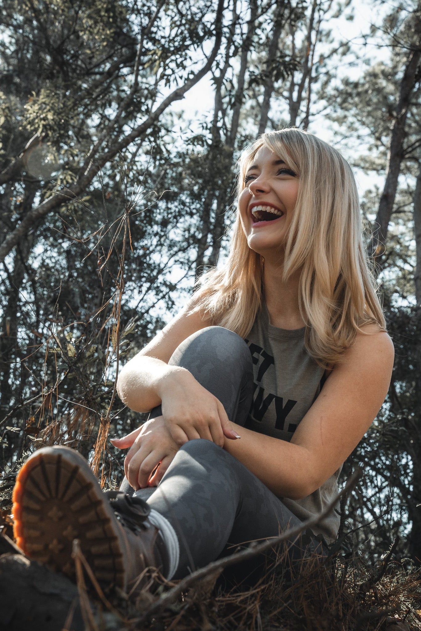 A woman with long blonde hair, in a sleeveless top and Born United Private Labels Midnight Camo Leggings, sits on the ground laughing in a sunlit forest surrounded by trees and plants, her joyful expression illuminated by sunlight filtering through the branches.