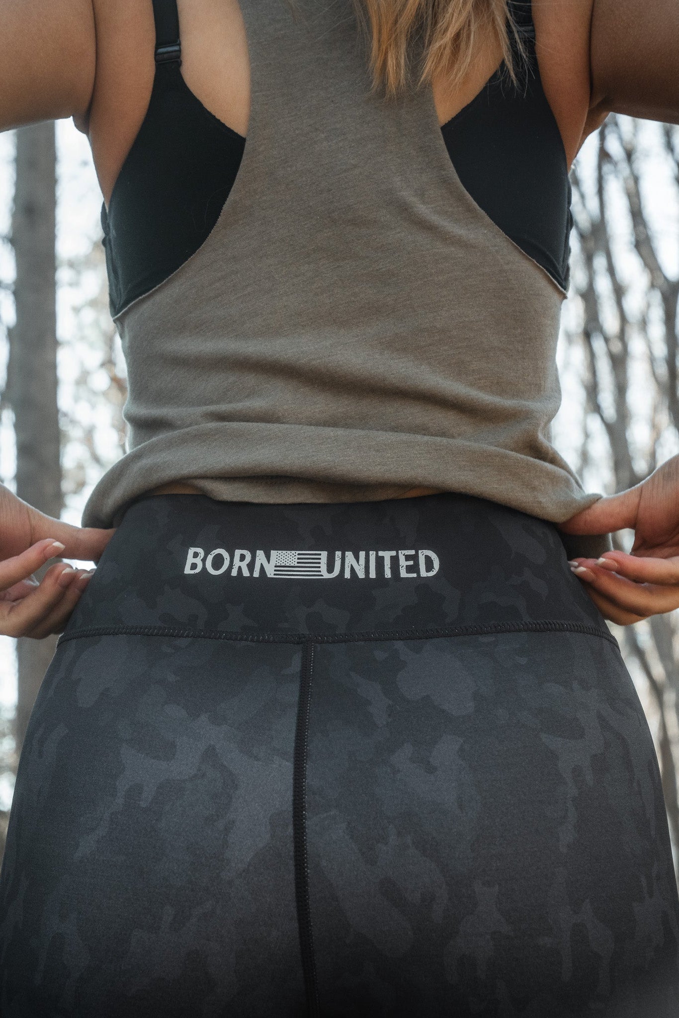 Amidst a natural setting, a person adjusts their Midnight Camo Leggings by Born United Private Label; the black leggings feature BORN UNITED on the waistband. They wear a gray sleeveless top surrounded by trees.