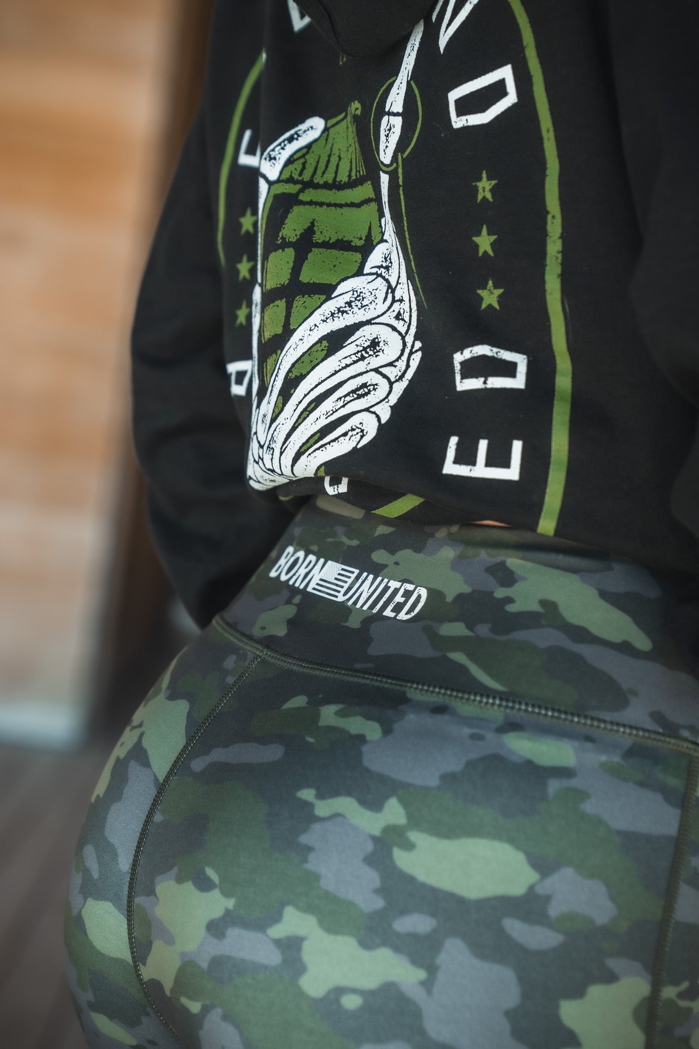 A person wears Combat Camo Leggings from Born United Private Label, featuring BORN UNITED on the waistband, paired with a black hoodie showcasing a graphic design. The focus is on the lower back and legs against a blurred wooden background.