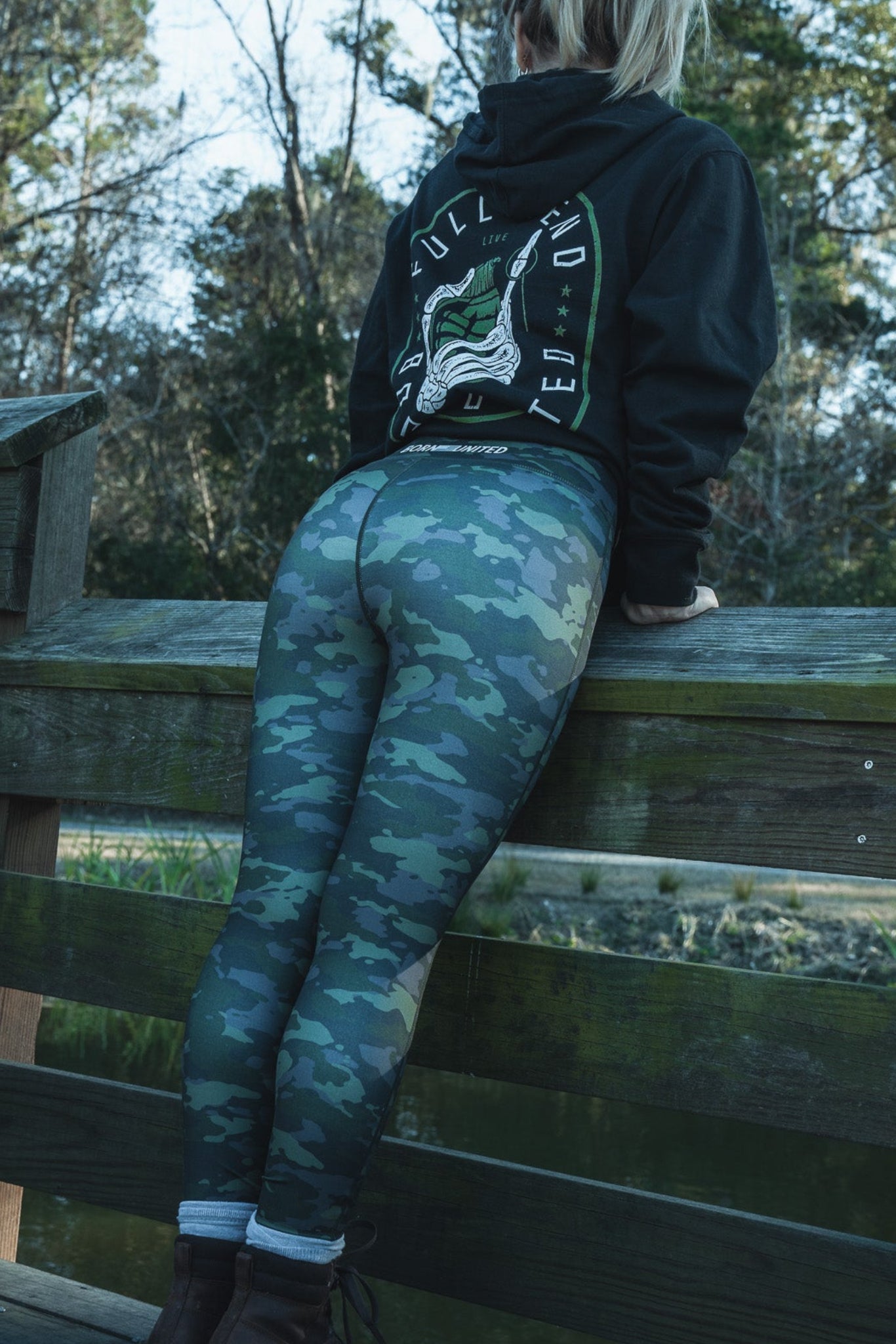 A person with blonde hair leans on a wooden bridge railing, gazing at the pond. Wearing their favorite black hoodie and Combat Camo Leggings by Born United Private Label, paired with brown boots, they stand surrounded by trees in the background.