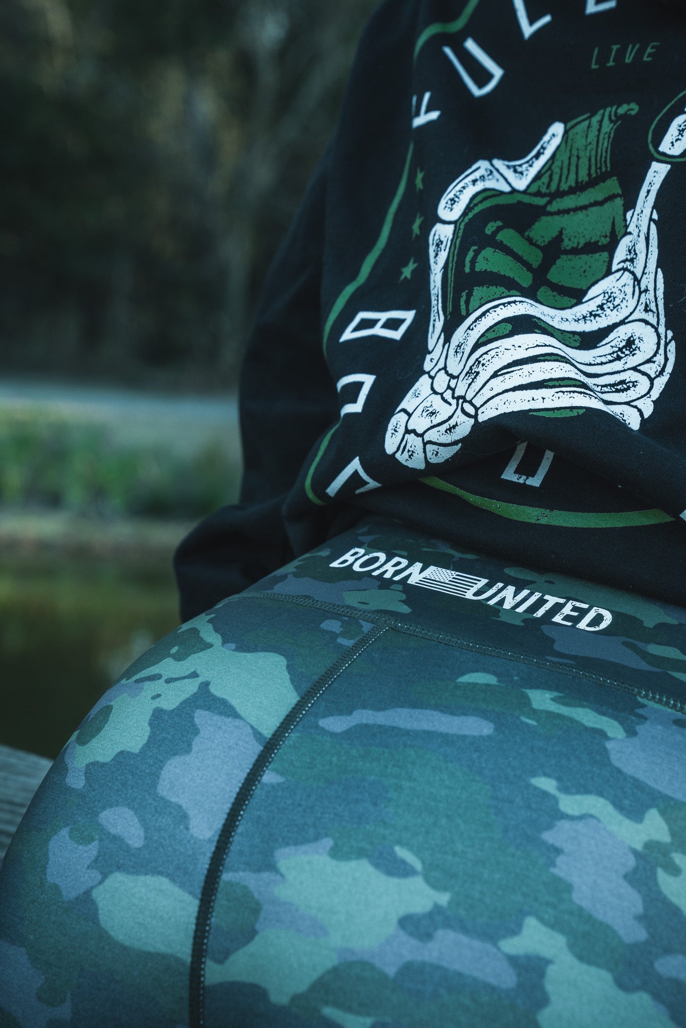 A person is seated outdoors wearing their favorite Combat Camo Leggings by Born United Private Label, along with a dark sweatshirt featuring a graphic of a hand making a peace sign.