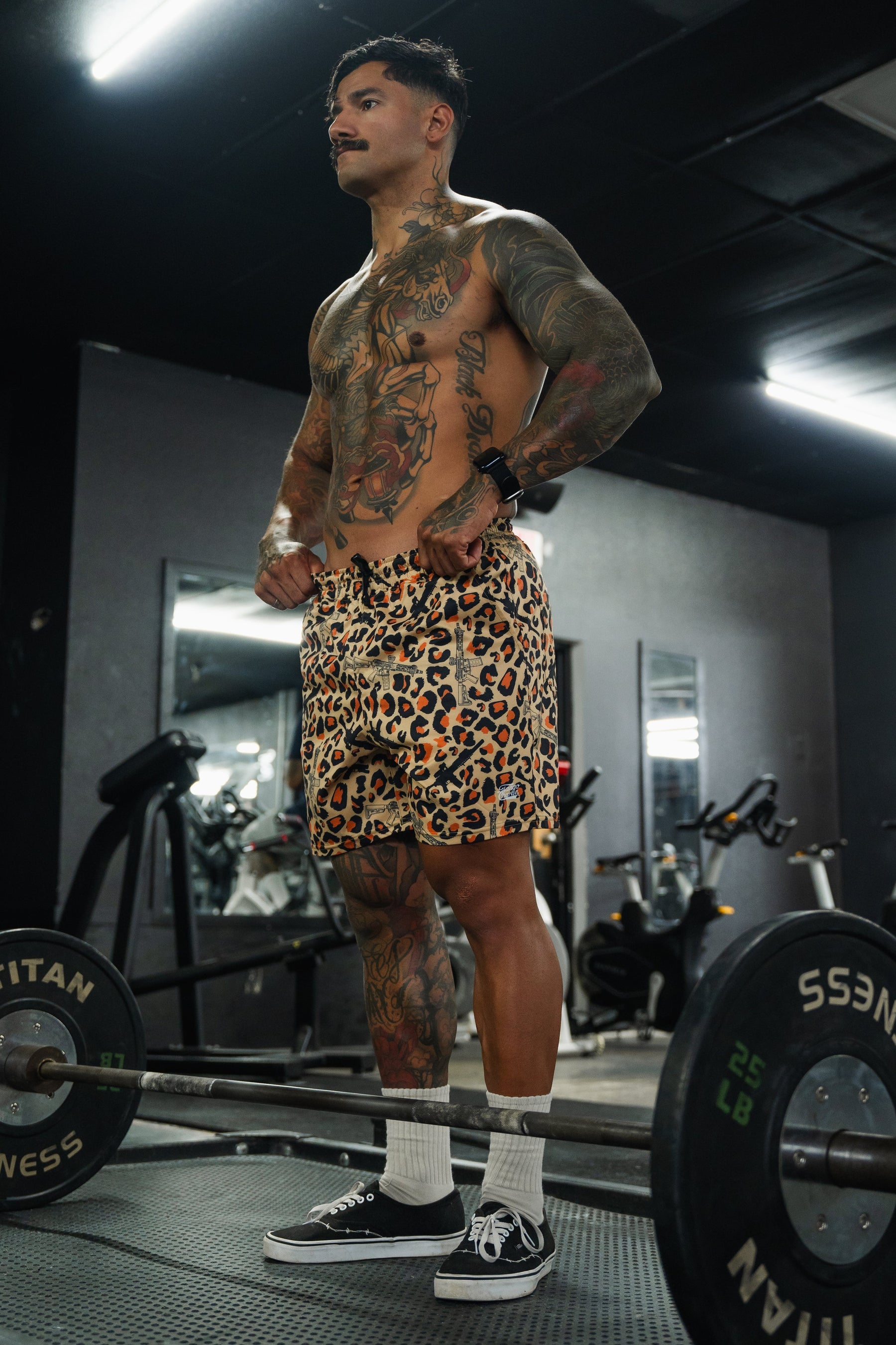 A muscular, tattooed man stands shirtless in a gym, wearing Tango Charlie Apparel Men's Performance Training Shorts | 5.5" - Loaded Leopard, paired with white socks and black sneakers. He poses by a barbell amid mirrors and workout equipment.