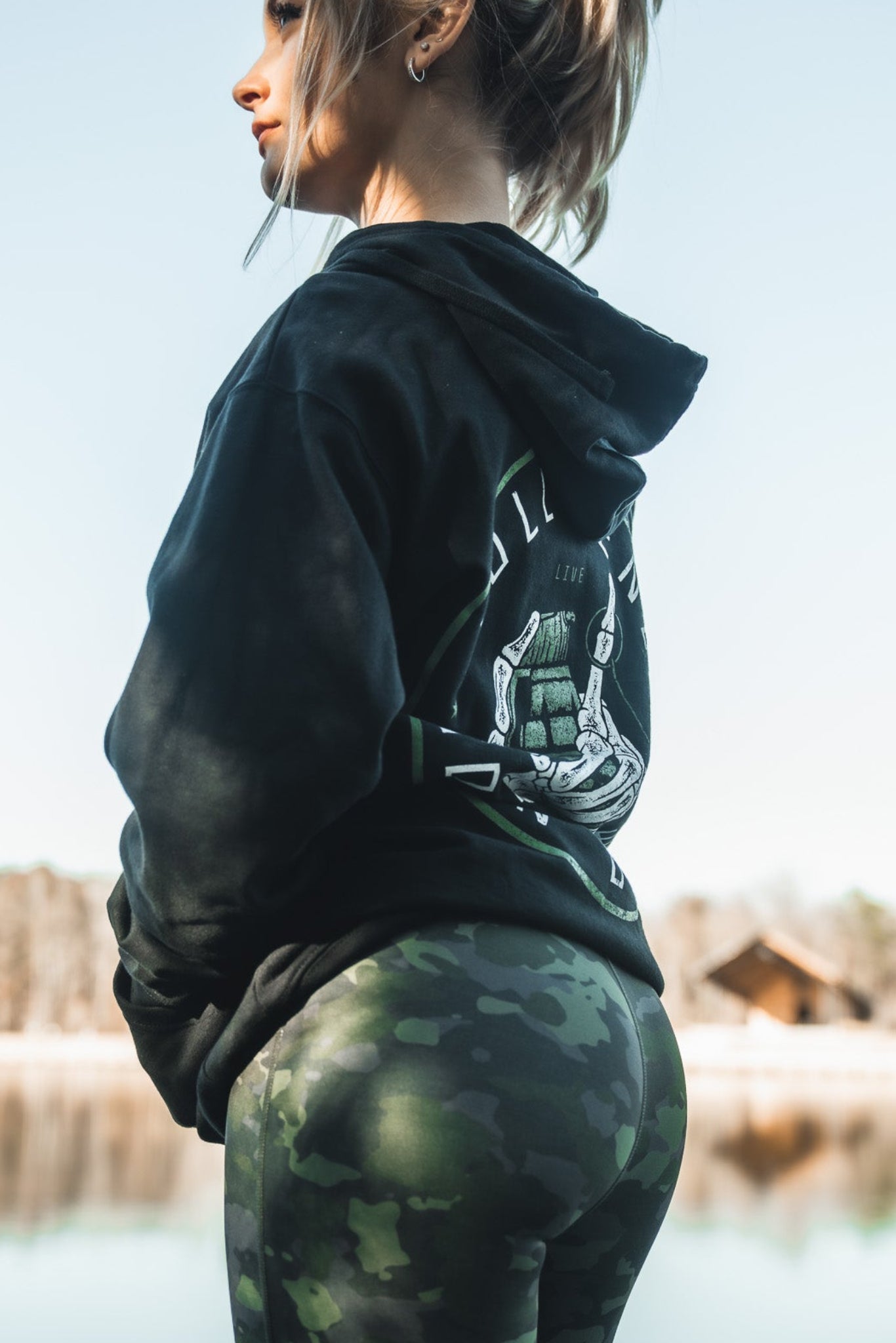 A woman wearing her favorite black hoodie and Combat Camo Leggings by Born United Private Label stands by a calm lake, gazing to the side. A wooden cabin and tree line frame the serene scene under a clear blue sky.