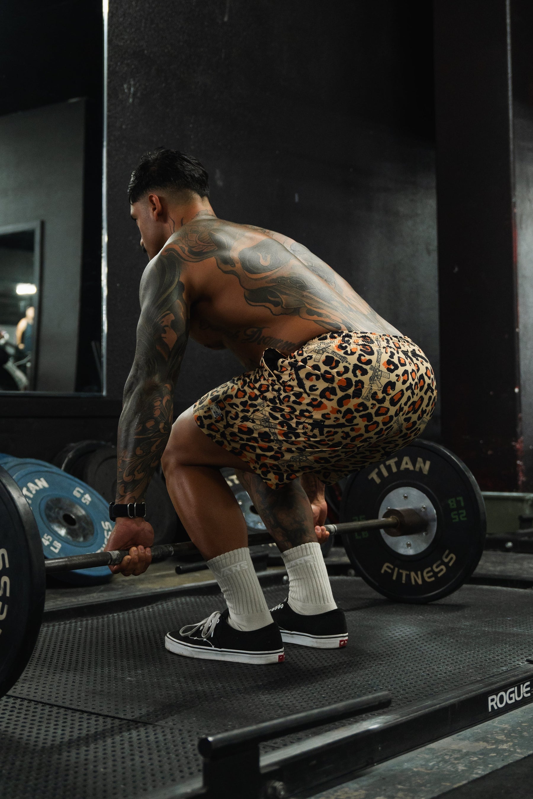 A muscular, tattooed individual wearing Tango Charlie Apparel Men's Performance Training Shorts | 5.5"- Loaded Leopard, white socks, and black sneakers squats in a gym, preparing to lift a barbell with weights.