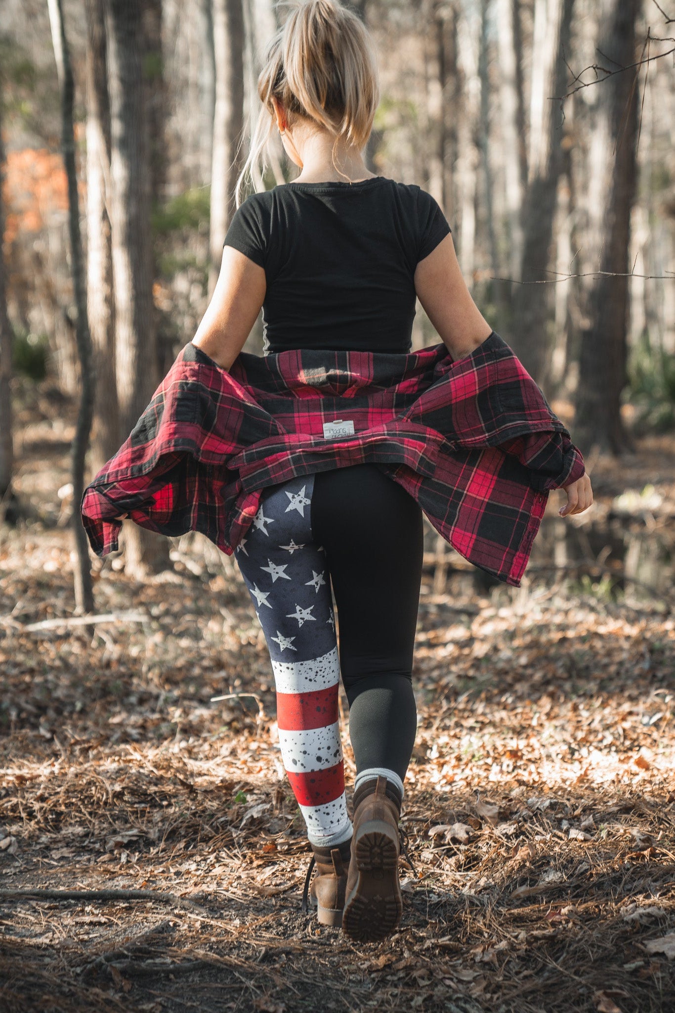 Strolling through the forest, a person with long hair sports a black shirt and Born United Private Labels American Savage Leggings, featuring stars and stripes on one leg. A red plaid jacket tied around their waist pairs perfectly with their brown boots as sunlight filters through the trees.