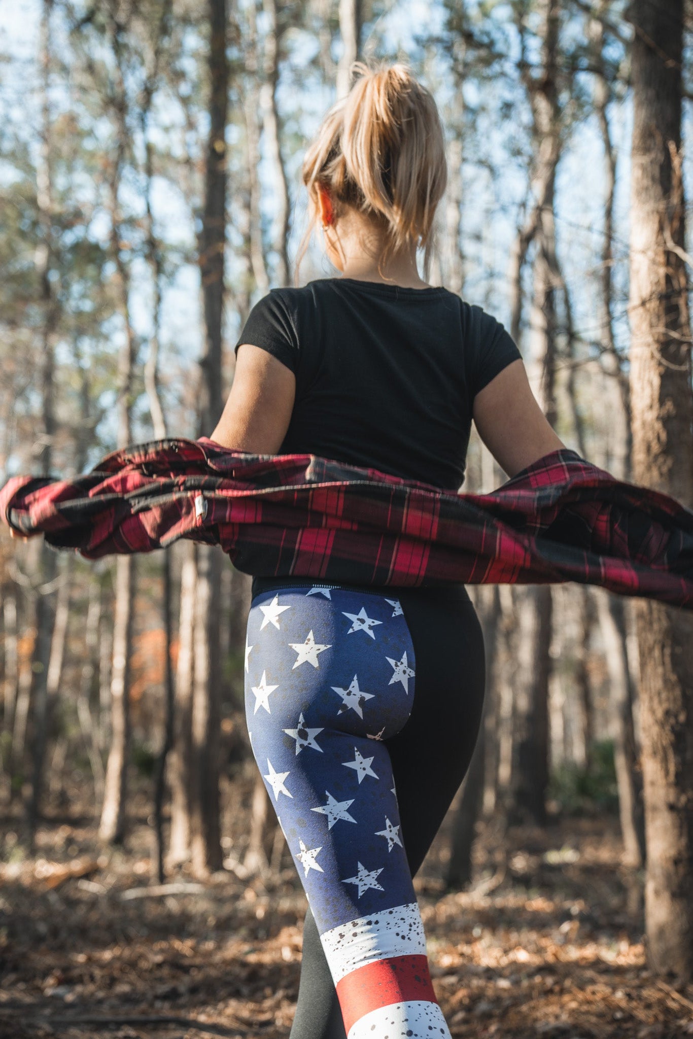 A woman strolls through the forest in American Savage Leggings by Born United Private Label, a black t-shirt, and clutching a red plaid shirt. Sunlight filters through the trees, creating a serene atmosphere.