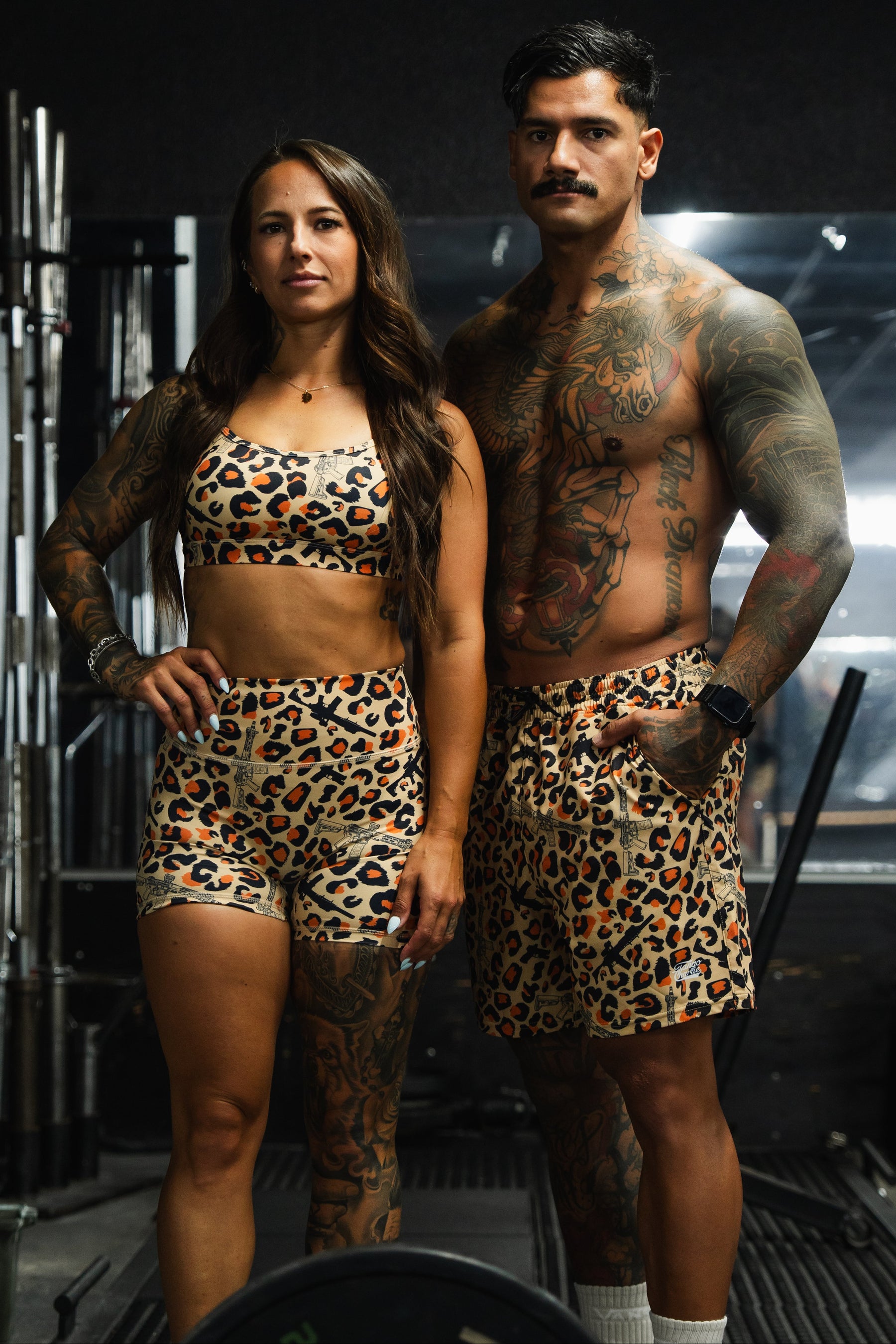 Two people at the gym show off tattoos side by side. The man wears Tango Charlie Apparel Men's Performance Training Shorts | 5.5"- Loaded Leopard, matching the woman's leopard print sports bra and shorts.