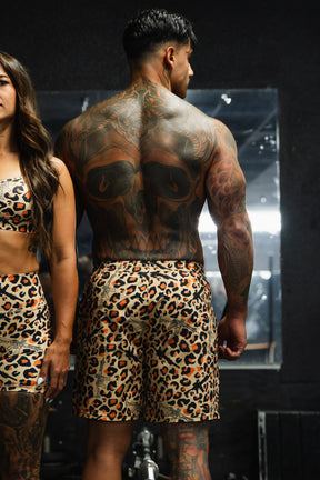 A man with a large skull tattoo on his back wears Tango Charlie Apparel's Men's Performance Training Shorts | 5.5"- Loaded Leopard, standing next to a woman in matching leopard print workout clothes in a gym setting.