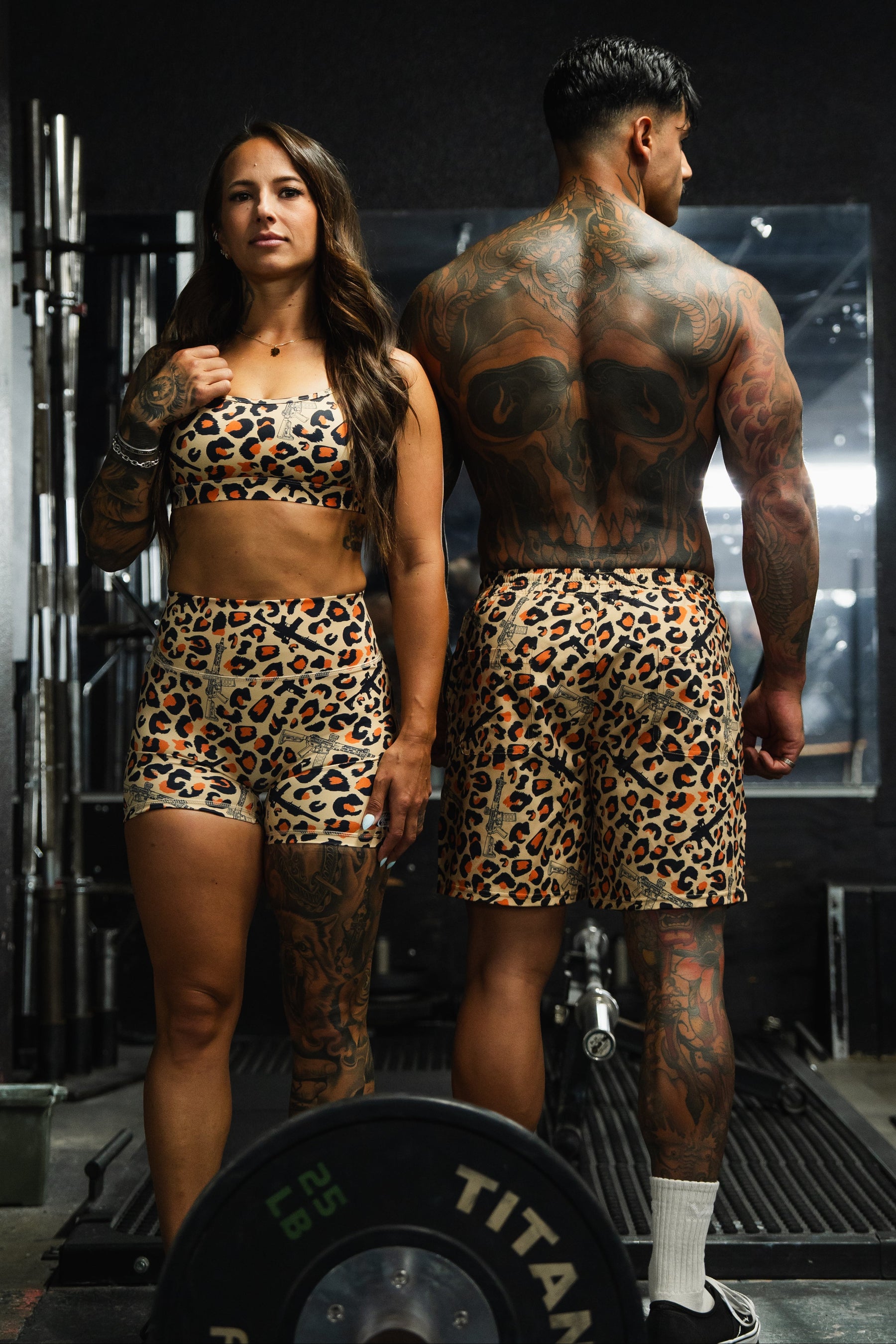 A woman wears Tango Charlie Apparel’s Women’s High-Waisted Seamless Front Compression Training Shorts in Loaded Leopard as she poses at the gym with a man; both sport matching leopard print outfits, surrounded by gym equipment.
