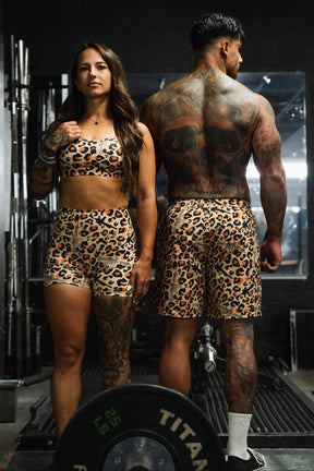A woman wears Tango Charlie Apparel’s Women’s High-Waisted Seamless Front Compression Training Shorts in Loaded Leopard as she poses at the gym with a man; both sport matching leopard print outfits, surrounded by gym equipment.