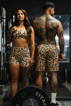 At the gym, a woman wears the Tango Charlie Apparel Y-Back Sports Bra in Loaded Leopard, paired with a man in matching leopard print athletic wear as they pose among weightlifting equipment, highlighting bold and stylish performance outfits.
