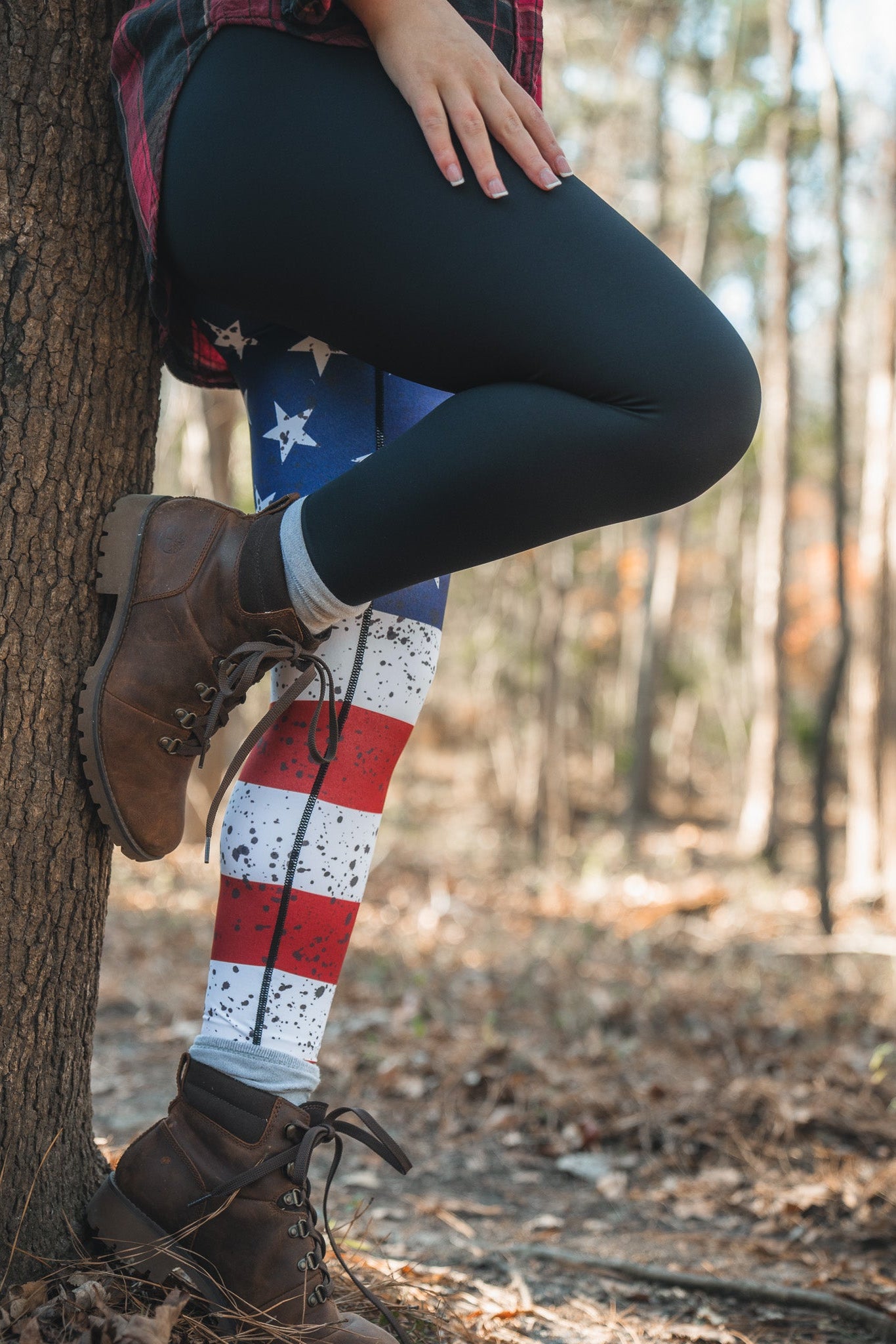 Amid fallen leaves and towering trees, a person stands by a tree, sporting American Savage Leggings from Born United Private Label beneath black pants. They wear a plaid shirt and brown boots, highlighting their patriotic style in the woods.
