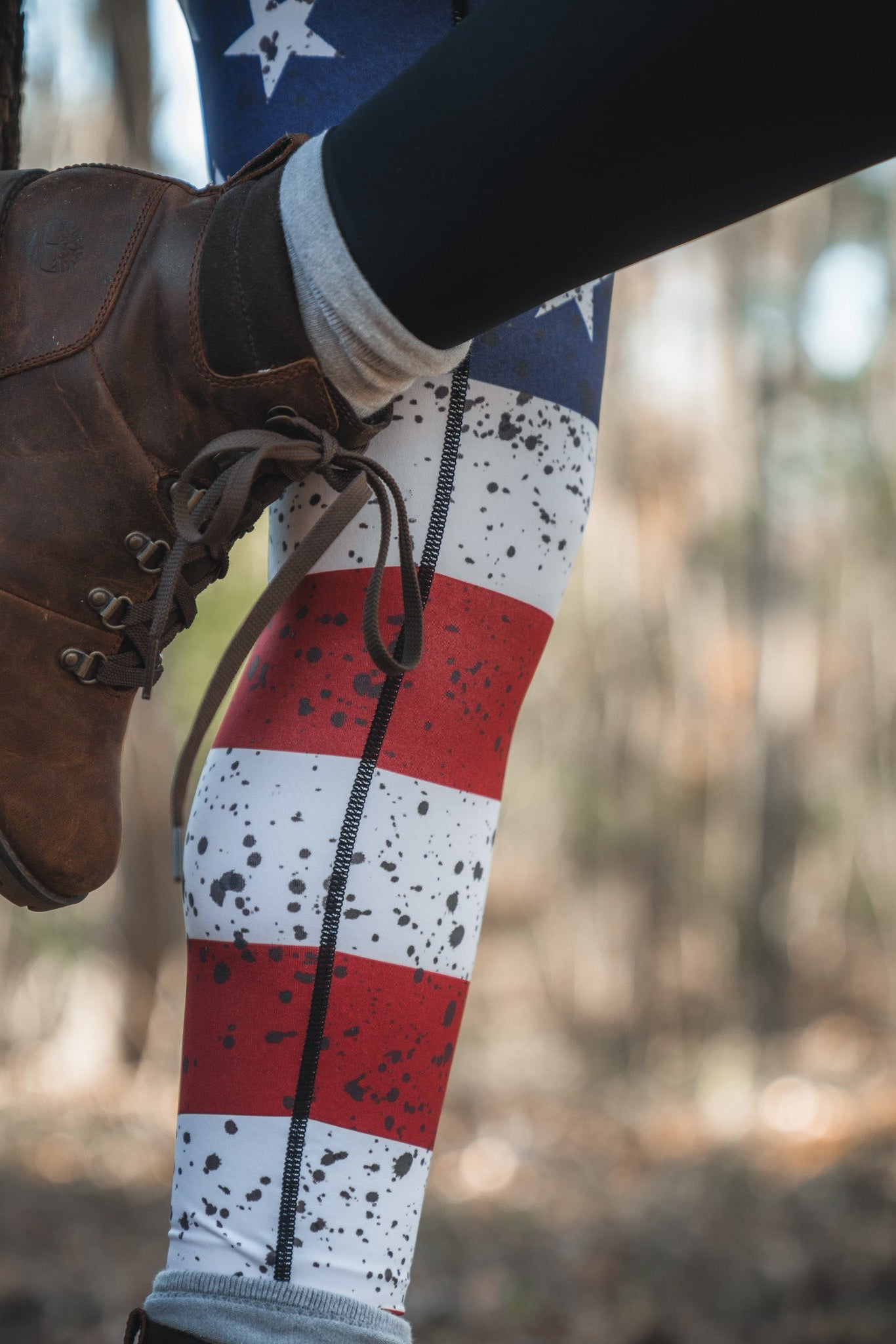 Wearing American Savage Leggings by Born United Private Label, featuring stars, stripes, and a splatter of red, white, and blue, a person in brown hiking boots rests their foot on a tree trunk within a lush forest.