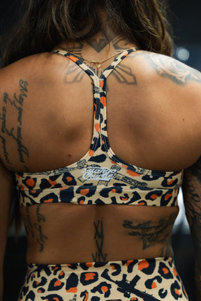 A person with long hair and several tattoos is photographed from behind, wearing the Tango Charlie Apparel Y-Back Sports Bra in Loaded Leopard.
