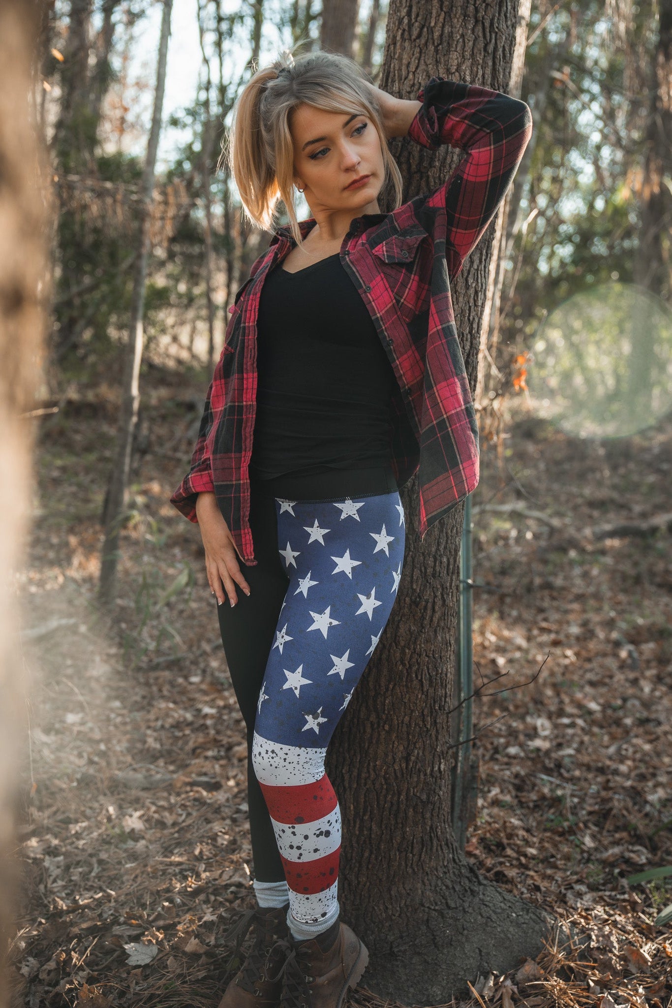 A woman wearing American Savage Leggings by Born United Private Label and a red plaid shirt leans against a sunlit forest tree, looking relaxed and contemplative with one hand behind her head and the other near her hip.