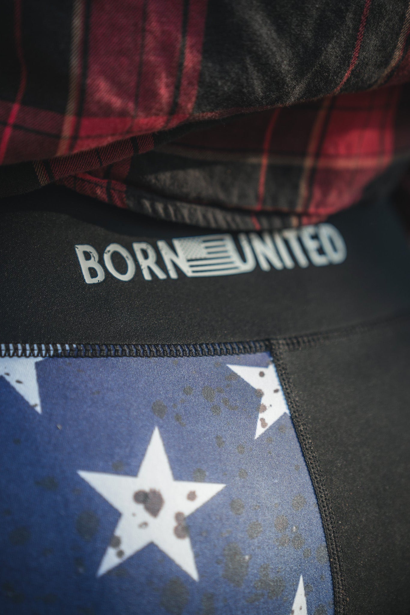 Close-up of someone wearing American Savage Leggings by Born United Private Label, featuring a stars and stripes pattern. BORN UNITED is emblazoned on the waistband with an American flag design in the letter R, paired with a red and black plaid shirt.