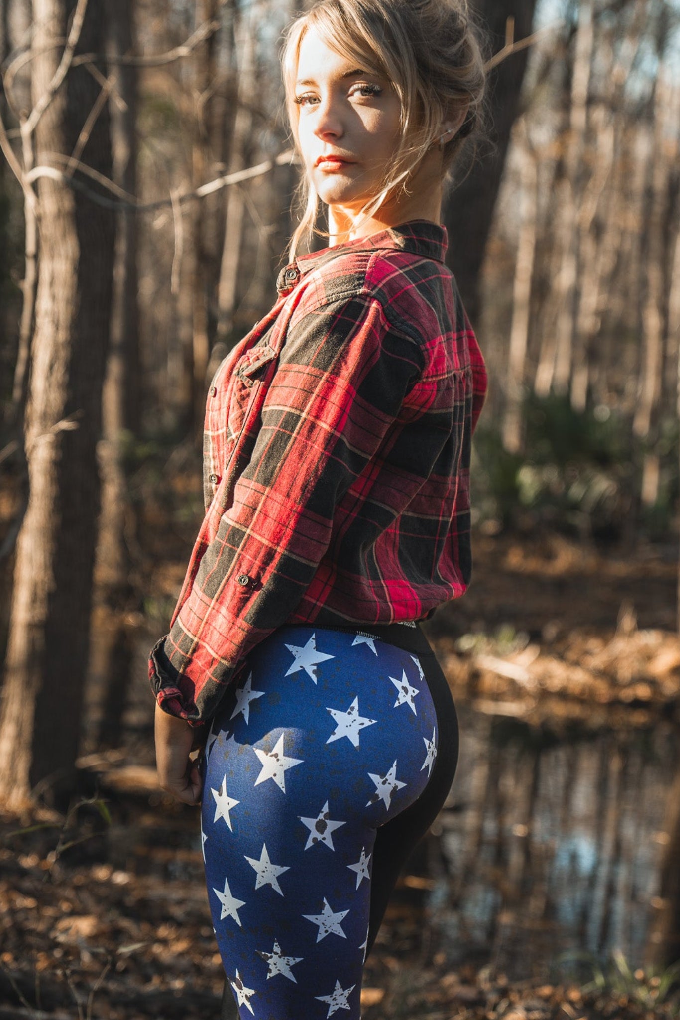 A person stands in a forest wearing Born United Private Labels red plaid shirt and patriotic American Savage Leggings, adorned with stars and stripes. Sunlight filters softly through the trees, casting shadows on the forest floor.
