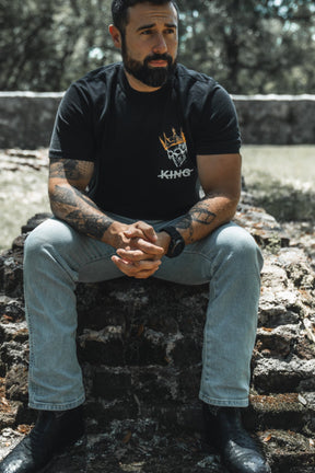 Man sitting on stone steps wearing a black t-shirt with a logo, light jeans, and black shoes.