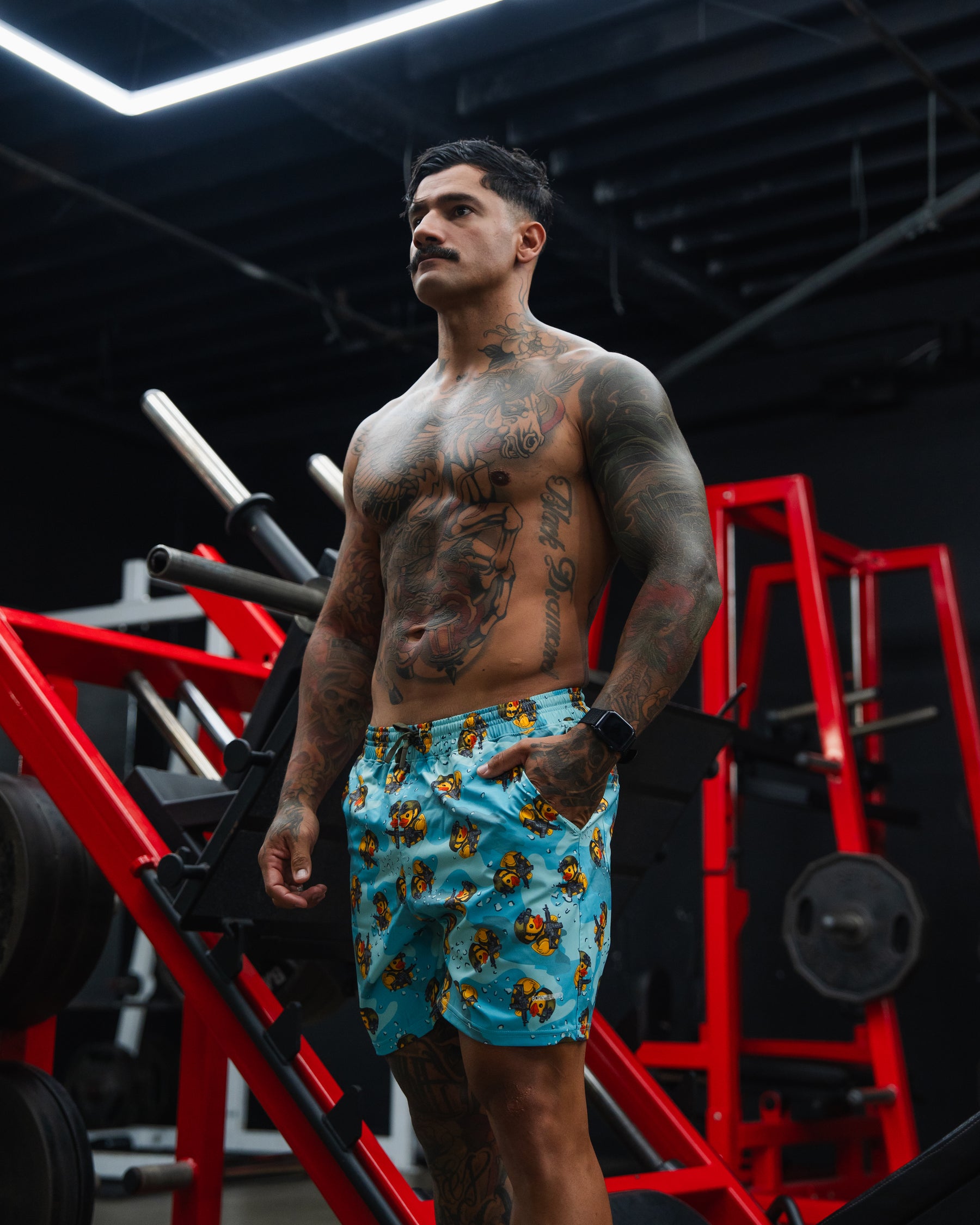 A muscular, tattooed man with a mustache stands shirtless in a gym, wearing Born United Private Label Men's Performance Hybrid Shorts 5.5" | TactiQuack. Red gym equipment and weight racks can be seen in the background.